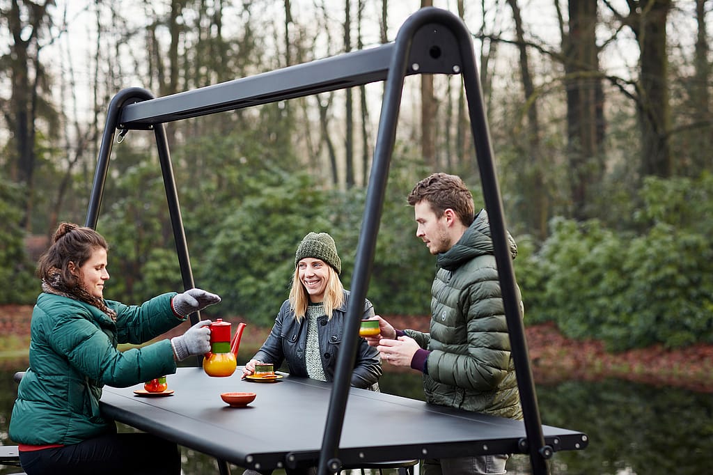 Drie mensen drinken thee aan een donkere AMAi hoge tafel met Tiki krukken op een houten dek naast een meer, omgeven door winterbomen in zacht zonlicht.