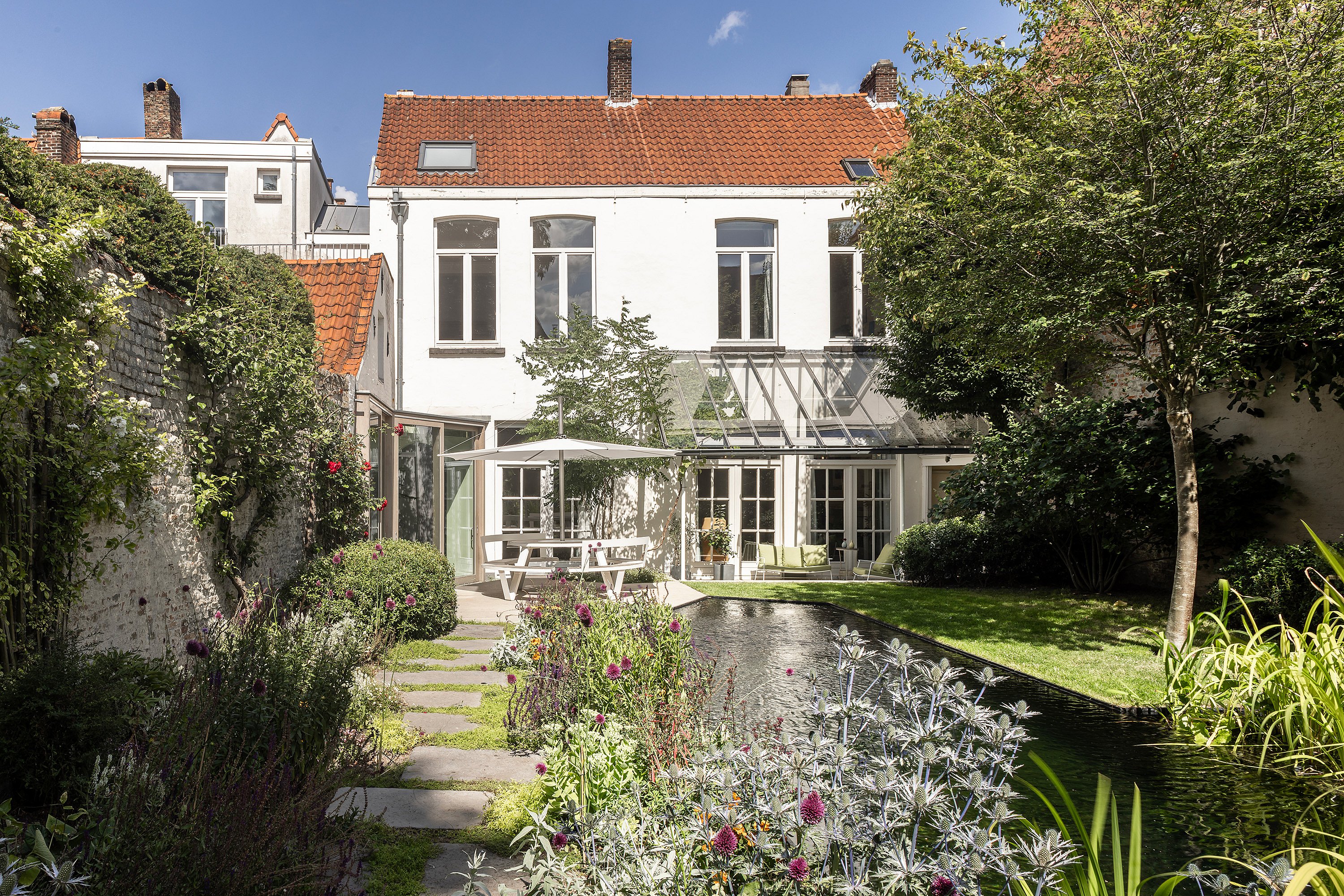 Rückansicht des Hauses mit Blick auf den Garten, mit dem Teich im Vordergrund und Terrassensitzen unter einem Sonnenschirm.