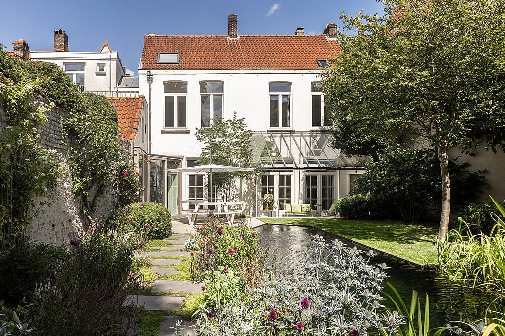 Rückansicht des Hauses mit Blick auf den Garten, mit dem Teich im Vordergrund und Terrassensitzen unter einem Sonnenschirm.