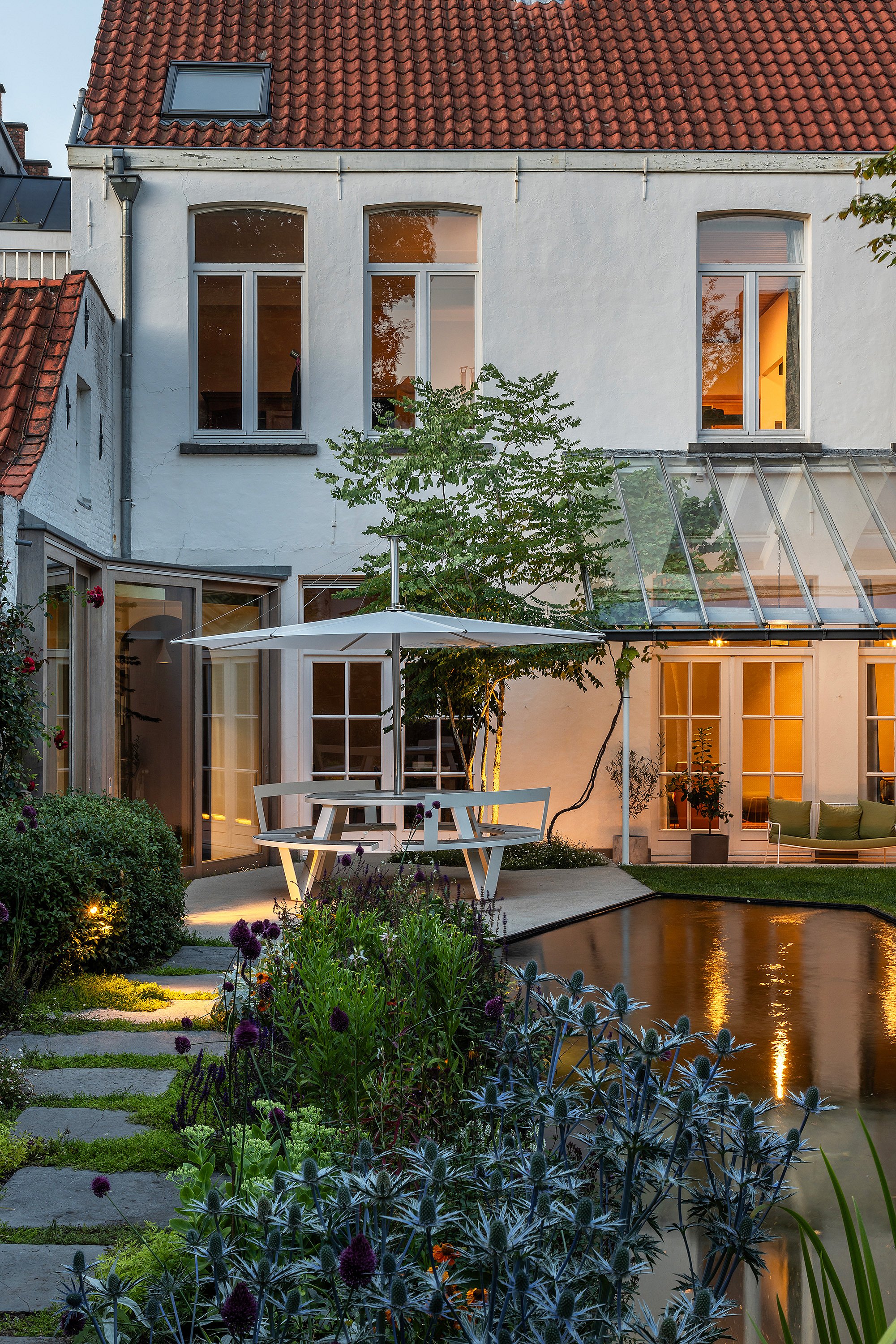 Nähere Abendansicht des Tisches und Sonnenschirms neben dem Teich, warmes Licht strömt aus dem Haus.