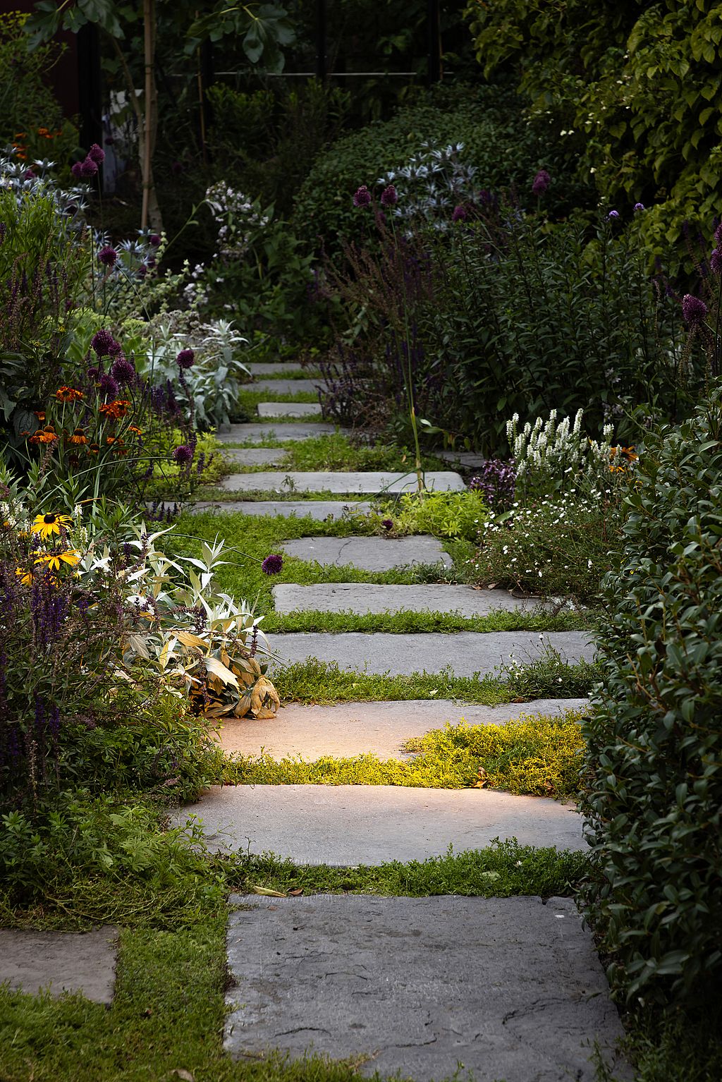 Abendlicher Blick auf den Gartenweg mit von Blumen gesäumten Trittsteinen und einem sanften Lichtbecken auf dem Gehweg.