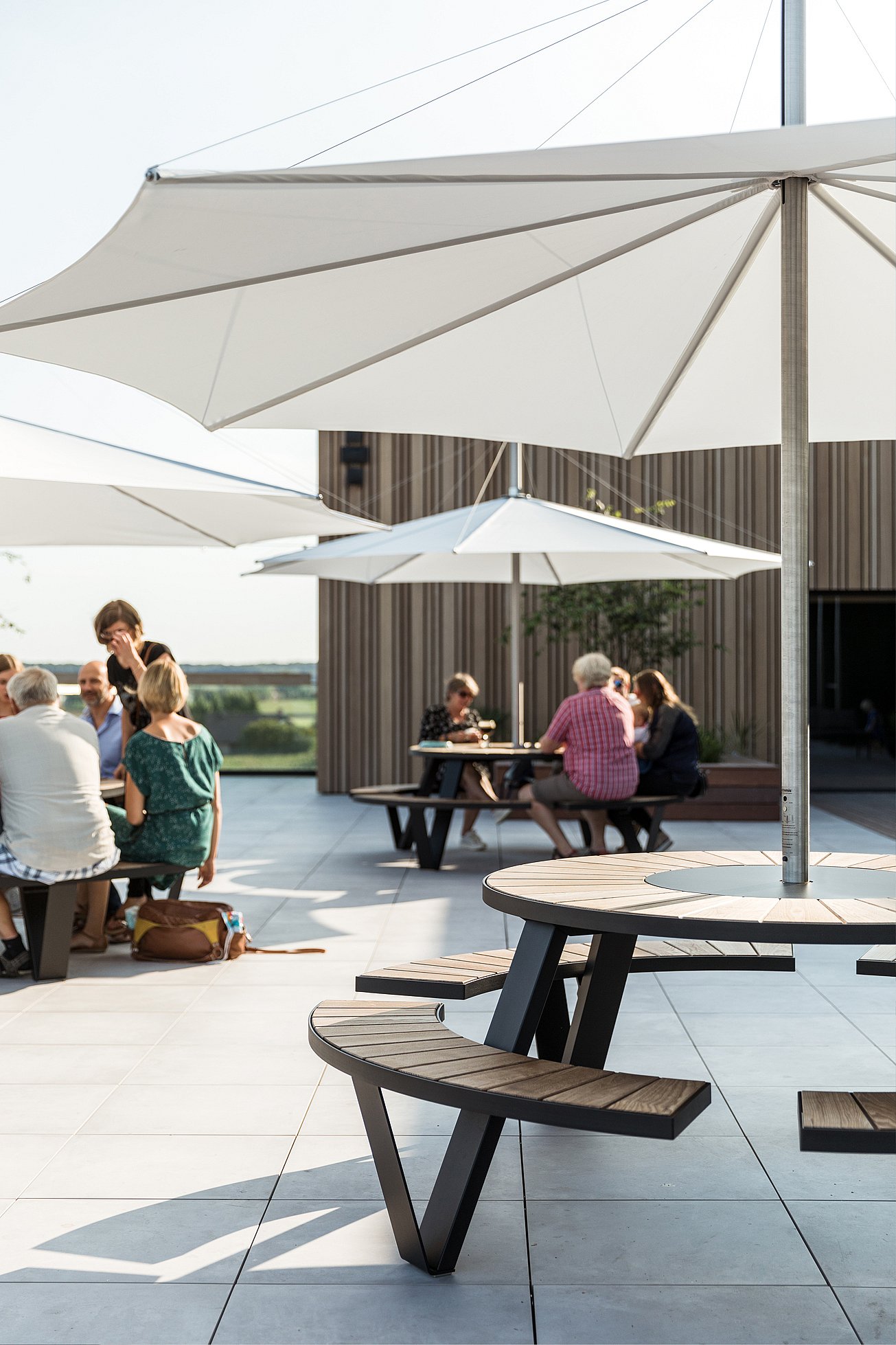 Des clients installés autour de tables en terrasse sur le toit, à l'ombre de grands parasols. On aperçoit les immeubles et la silhouette de la ville en arrière-plan.