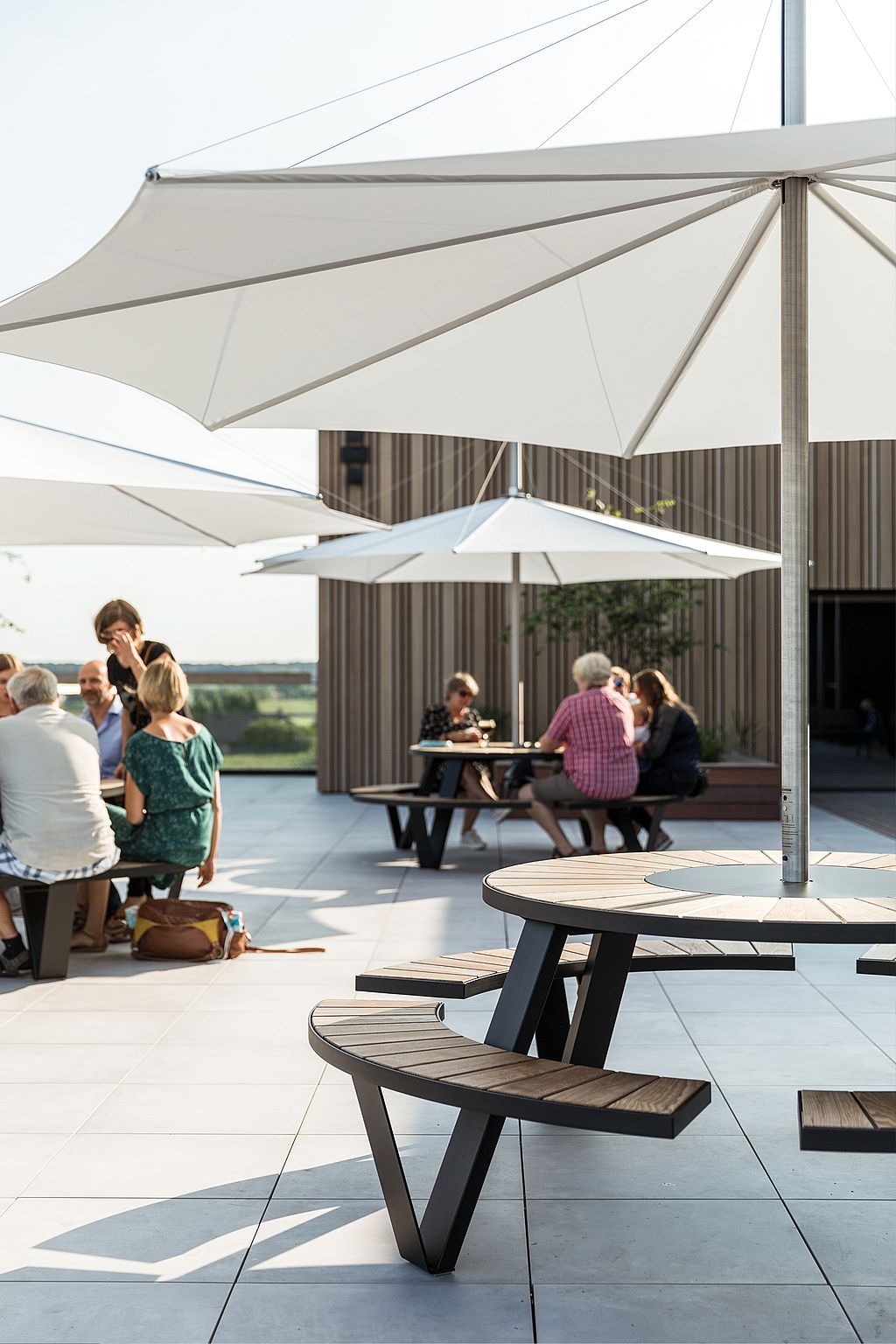 Des clients installés autour de tables en terrasse sur le toit, à l'ombre de grands parasols. On aperçoit les immeubles et la silhouette de la ville en arrière-plan.