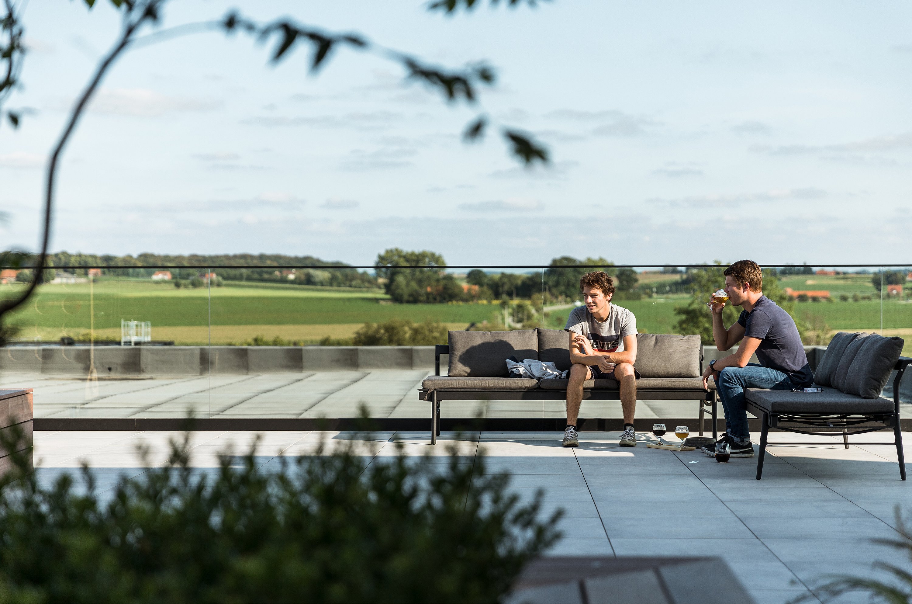 Deux jeunes hommes, assis sur un Sol+Luna, profitent de la vue depuis la terrasse sur le toit, dotée d'une balustrade en verre. L'un d'eux sirote un verre tandis que l'autre se détend près d'une table basse où sont posés d'autres boissons.