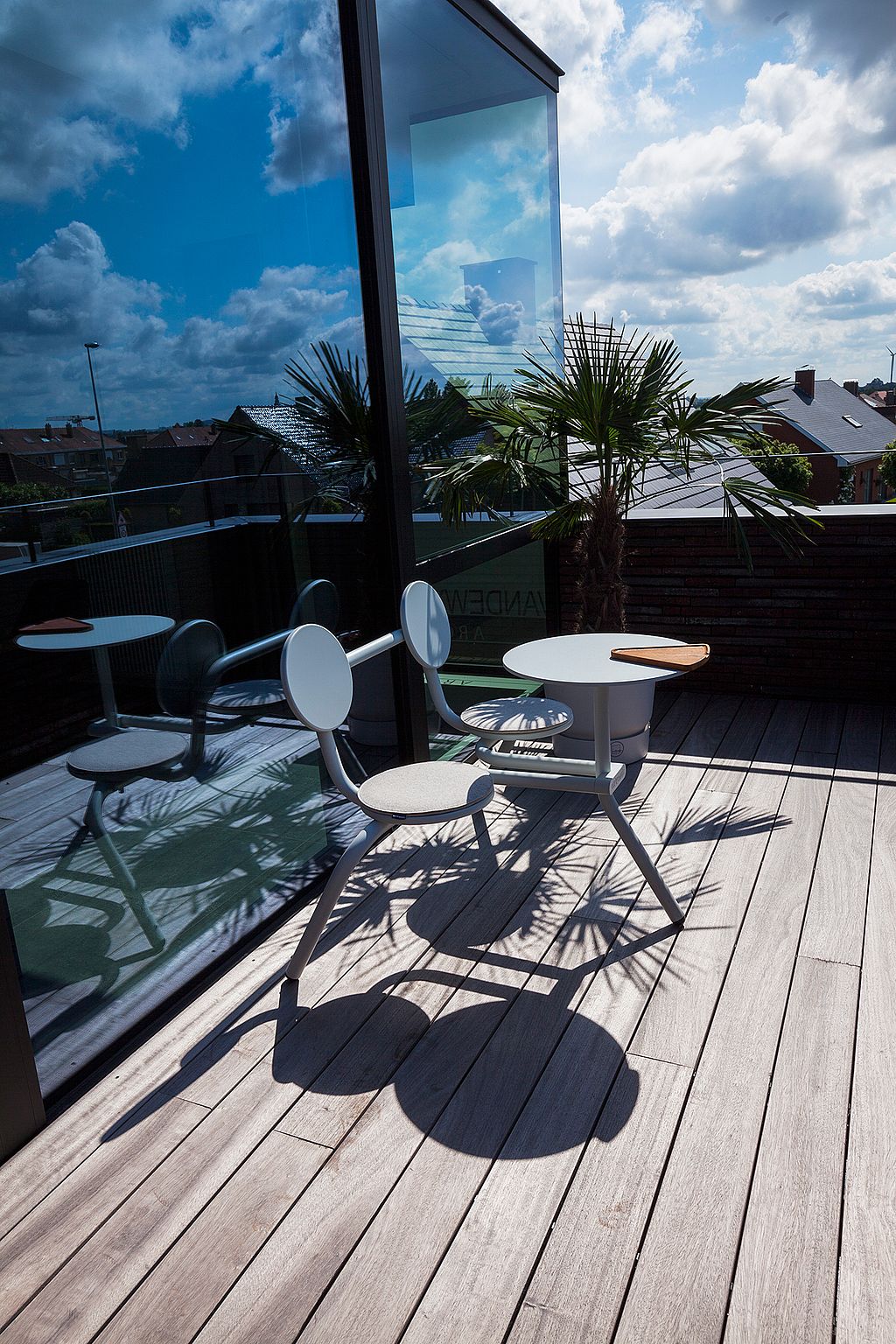 Witte Bistroo-tafel en -stoel voor twee personen op een zonnig dakterras, weerspiegeld in een kamerhoge glazen wand met daken en wolken op de achtergrond.