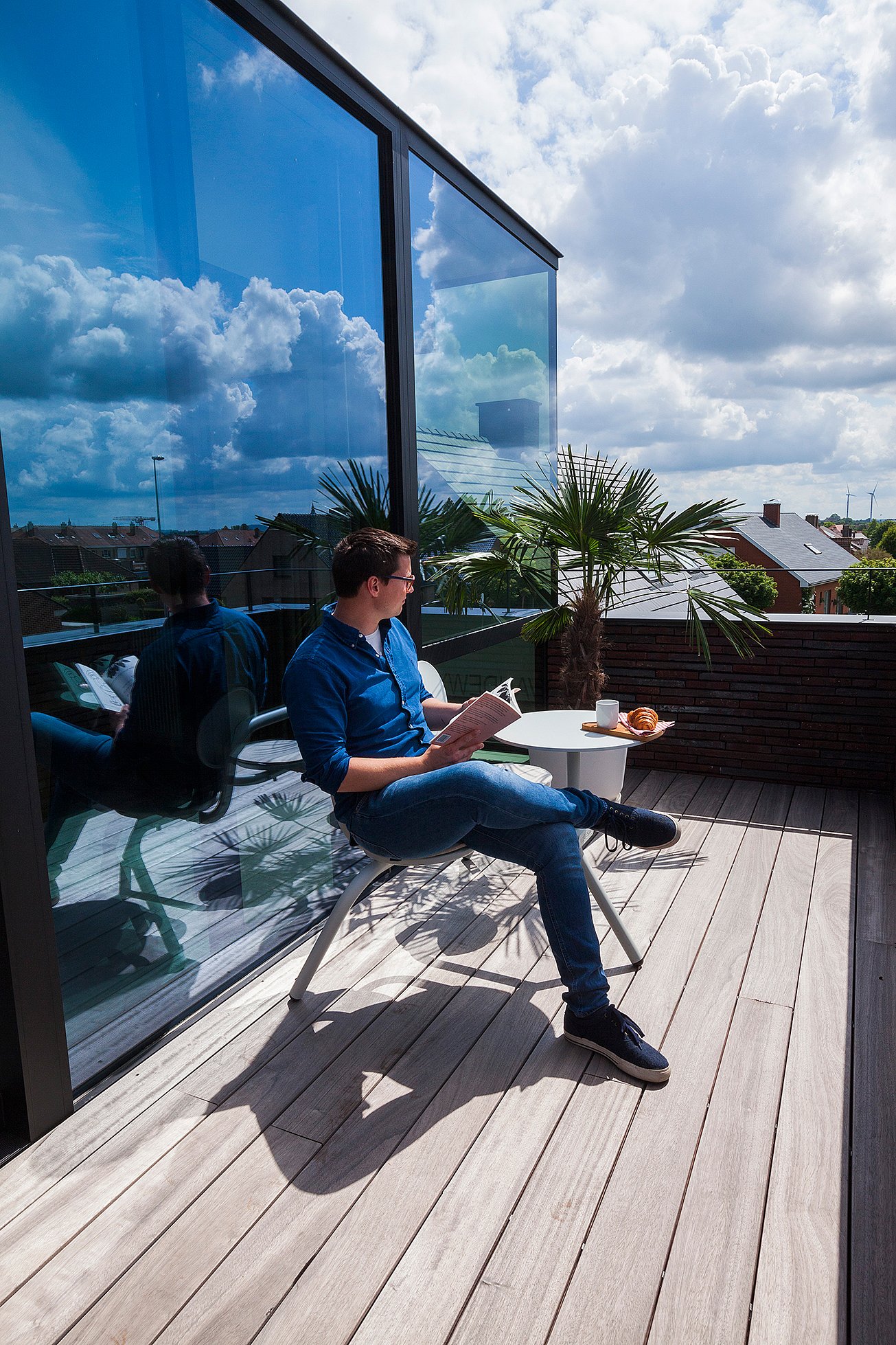 Een man ontspant op een dakterras, lezend aan een Bistroo-tafel met koffie en een croissant, naast een potpalm en een reflecterende glazen gevel.