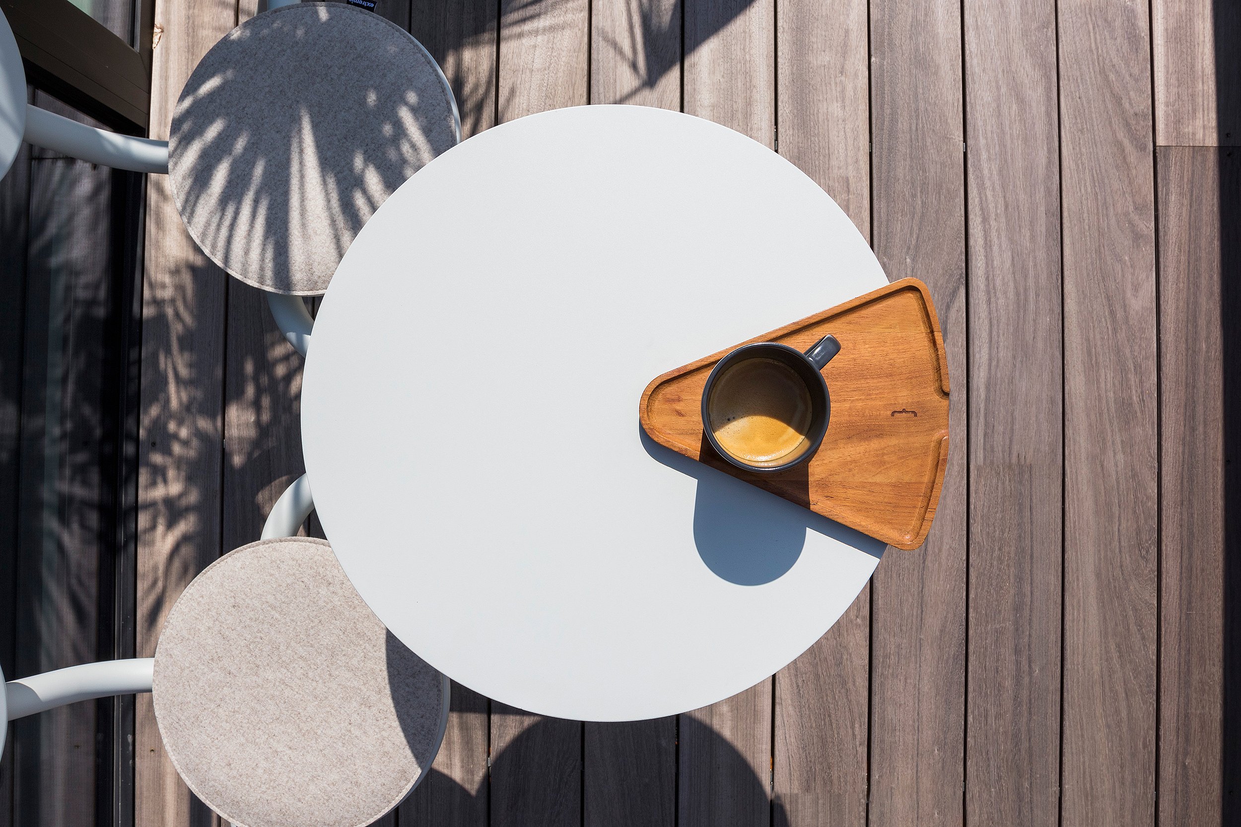 Overzicht van een rond wit Bistroo-tafelblad met een kop koffie op een driehoekig houten dienblad, omringd door twee gebogen stoelen op een houten terras.
