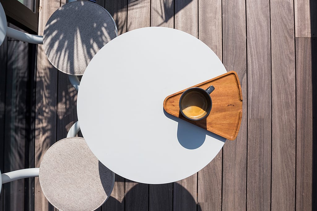 Overzicht van een rond wit Bistroo-tafelblad met een kop koffie op een driehoekig houten dienblad, omringd door twee gebogen stoelen op een houten terras.