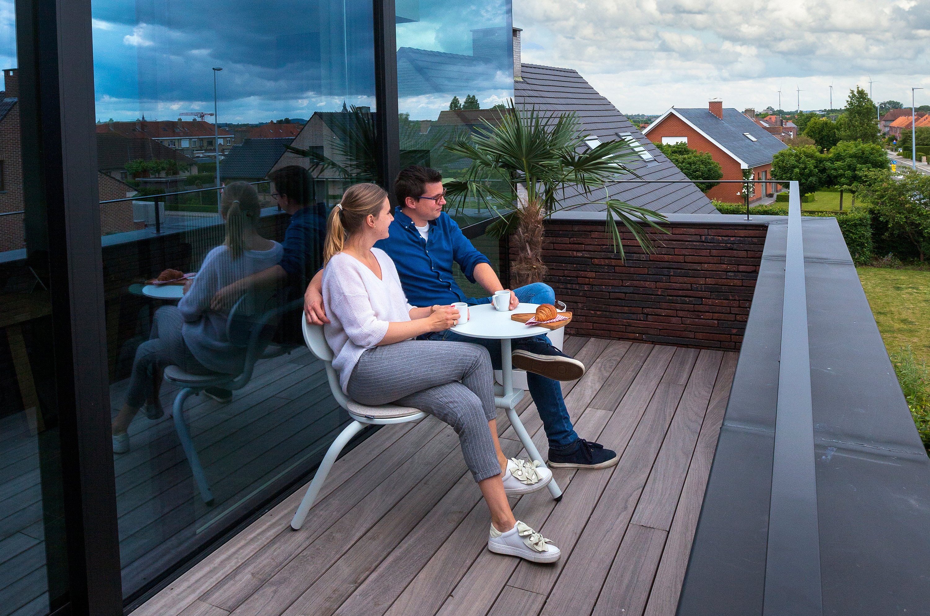 Couple enjoying breakfast on a modern rooftop balcony, seated side by side at a small round Bistroo table with coffee and croissants, next to a potted palm and glass façade.
