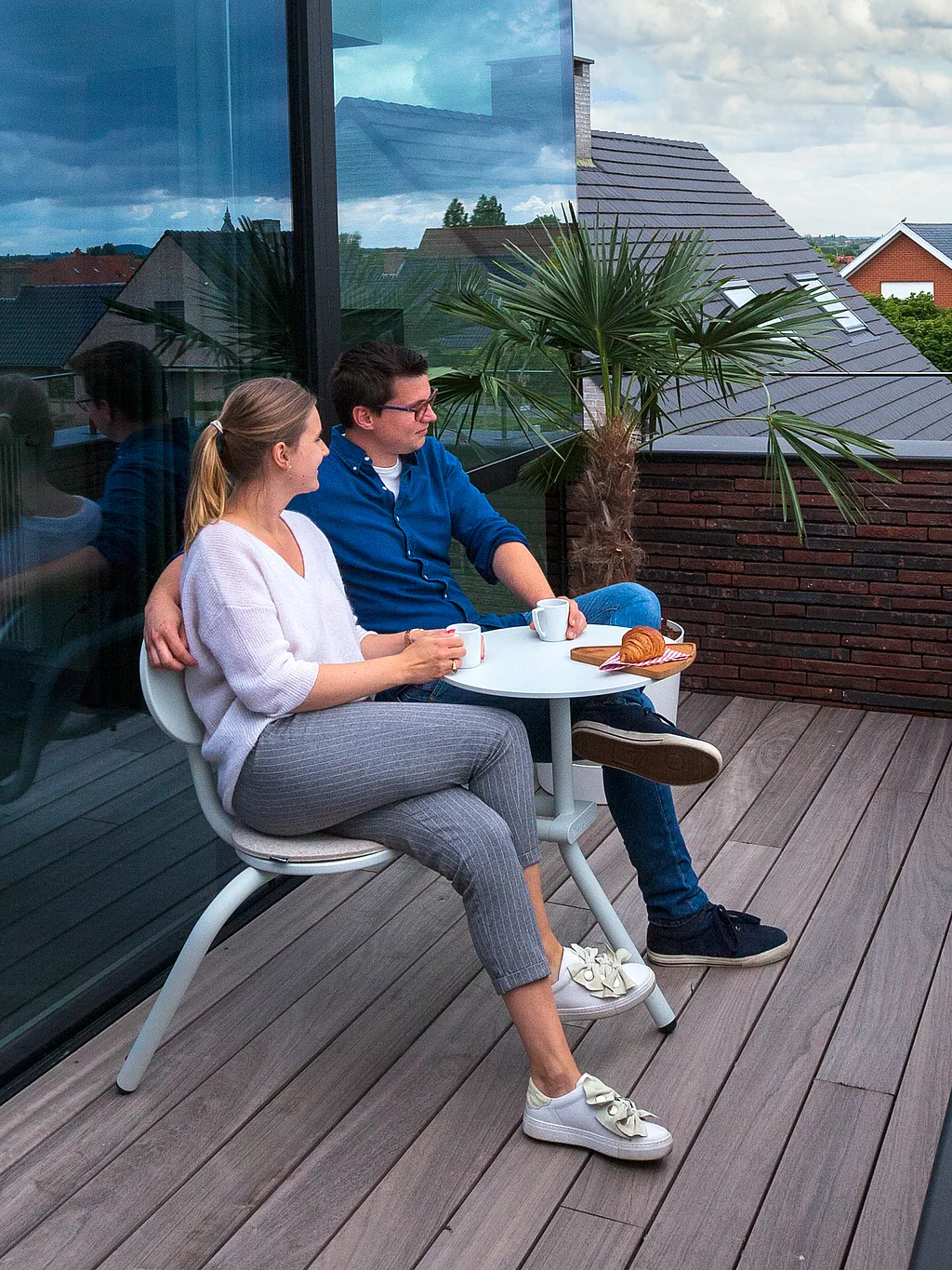 Een stel geniet van hun ontbijt op een modern dakterras, naast elkaar zittend aan een kleine ronde Bistroo-tafel met koffie en croissants, naast een potpalm en een glazen gevel.