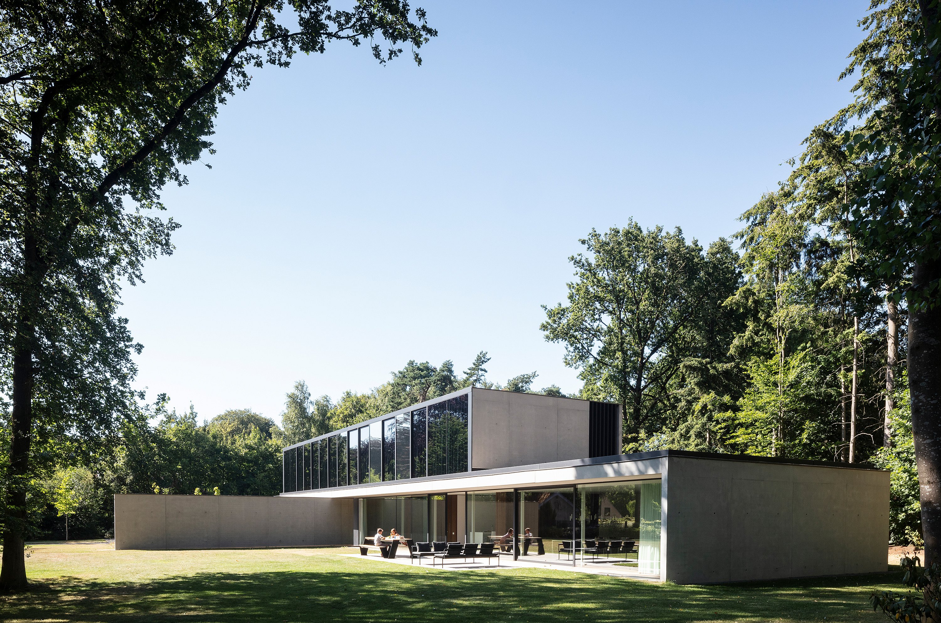 Villa contemporaine en béton et verre, nichée dans un paysage boisé, avec de grandes baies vitrées coulissantes et un long volume horizontal à l'étage. Sur la terrasse, le salon de jardin sombre Sol+Luna et la table de pique-nique Hopper sont disposés sur le patio donnant sur la terrasse, sous de grands arbres.