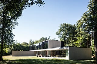 Villa contemporaine en béton et verre, nichée dans un paysage boisé, avec de grandes baies vitrées coulissantes et un long volume horizontal à l'étage. Sur la terrasse, le salon de jardin sombre Sol+Luna et la table de pique-nique Hopper sont disposés sur le patio donnant sur la terrasse, sous de grands arbres.