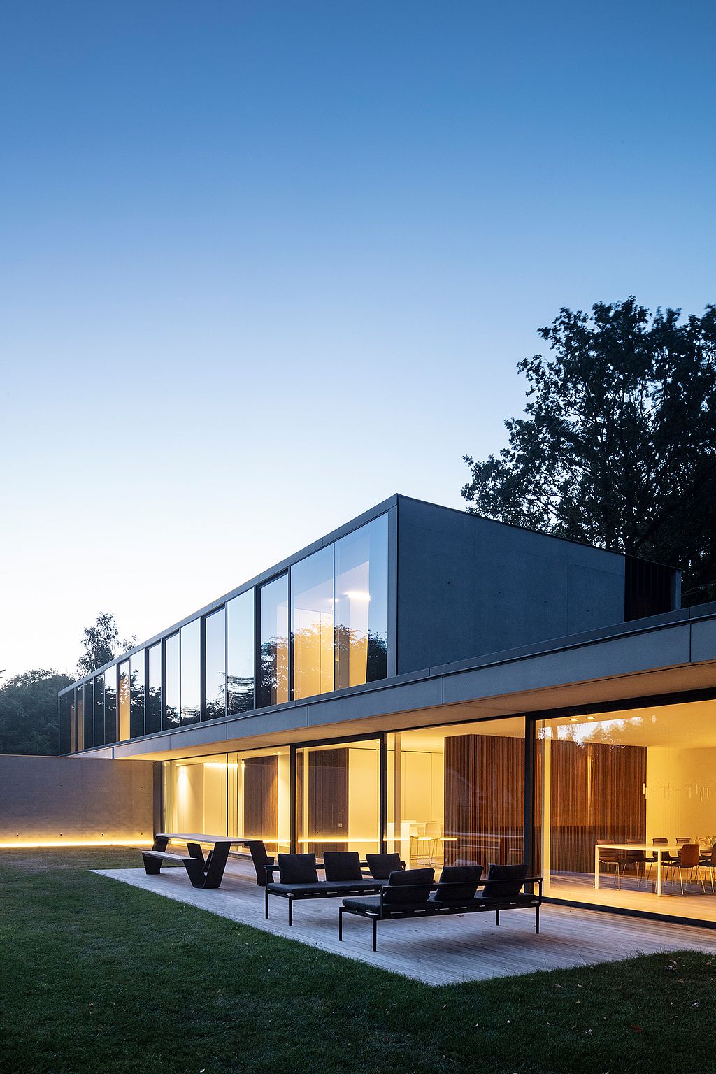 Maison moderne et minimaliste avec de grandes baies vitrées illuminées au crépuscule. Devant la maison, un salon de jardin foncé Sol+Luna et une table de pique-nique Hopper sont installés sur une terrasse en bois bordée de pelouse.