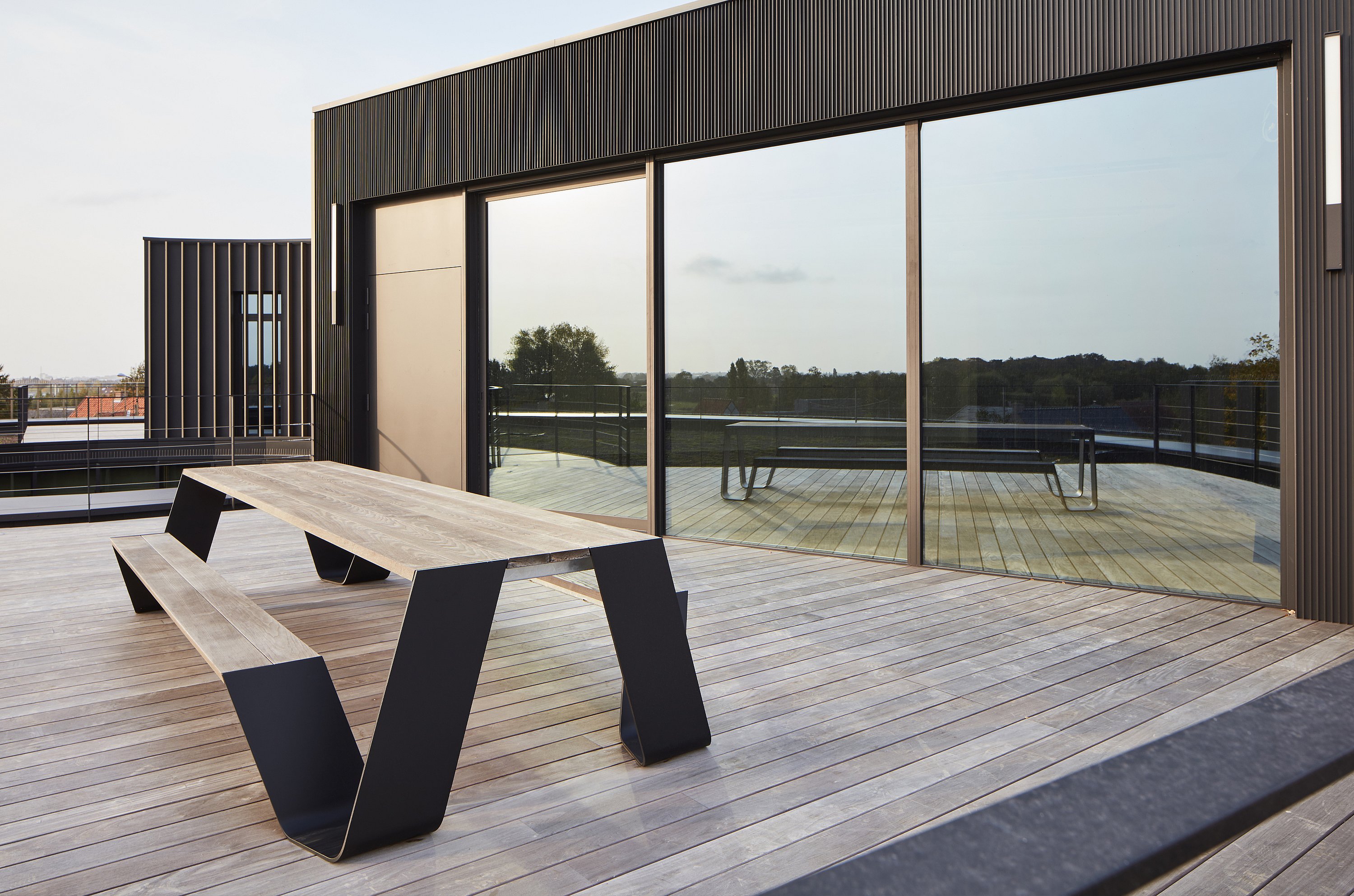Op een dakterras naast grote reflecterende ramen staat een eigen houten hoptafel.