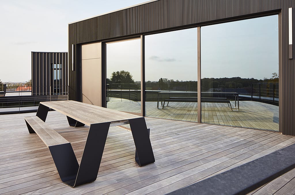 Op een dakterras naast grote reflecterende ramen staat een eigen houten hoptafel.