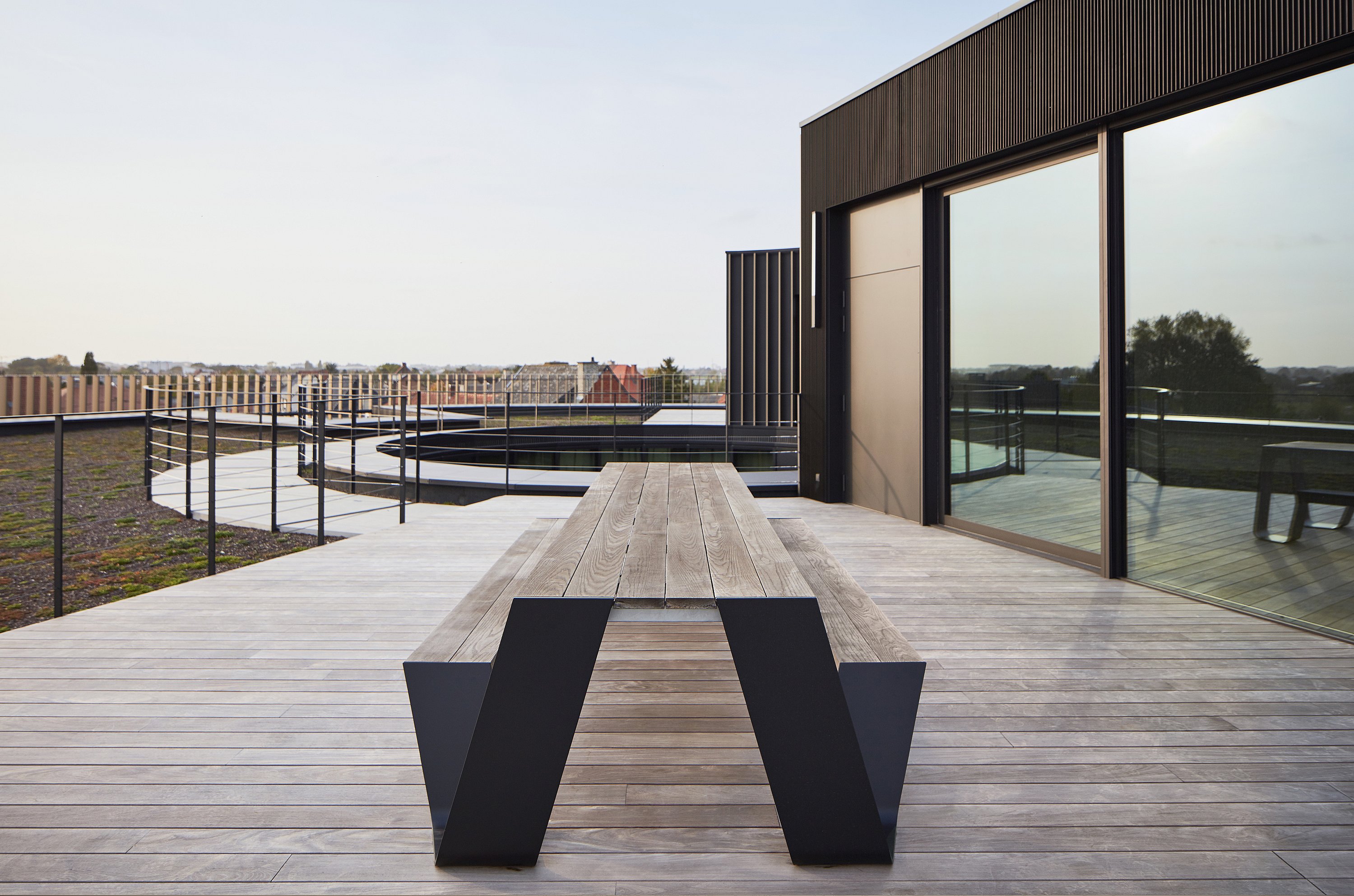 Achteraanzicht van een lange Hopper picknicktafel op een dakterras, met uitzicht op de leuningen en terrassen van het ronde kantoorgebouw.