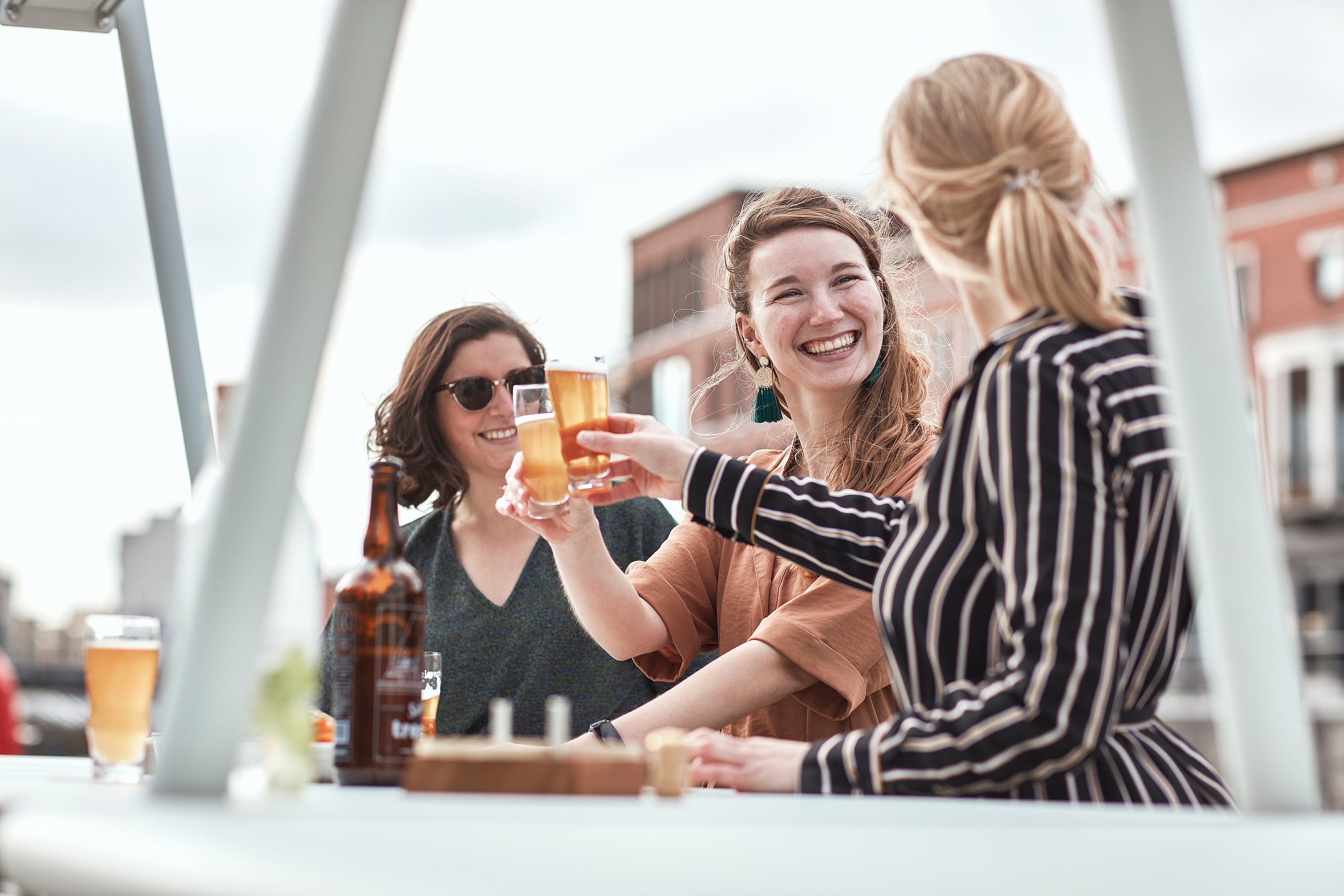 Three women clink beer glasses at the outdoor table, framed by the light grey structure and city buildings behind.