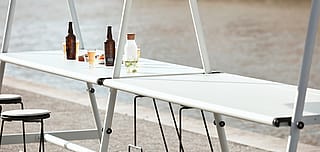 Long, light grey outdoor table beside the river with stools, bottles and glasses set out for a meal.