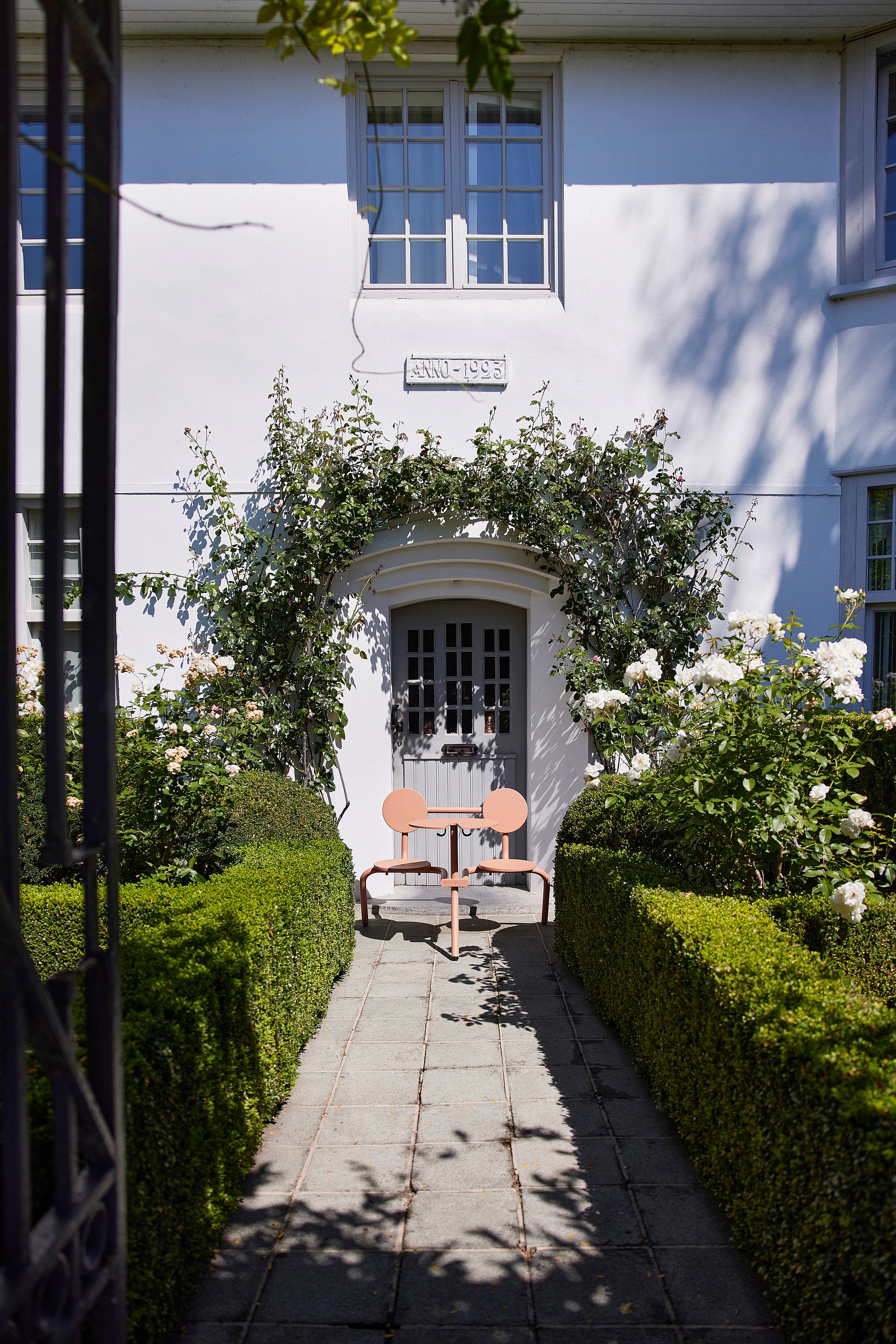 Ensemble Bistroo vide placé sur un chemin pavé dans un jardin entre des haies taillées, face à la porte d'une maison blanche recouverte de roses.