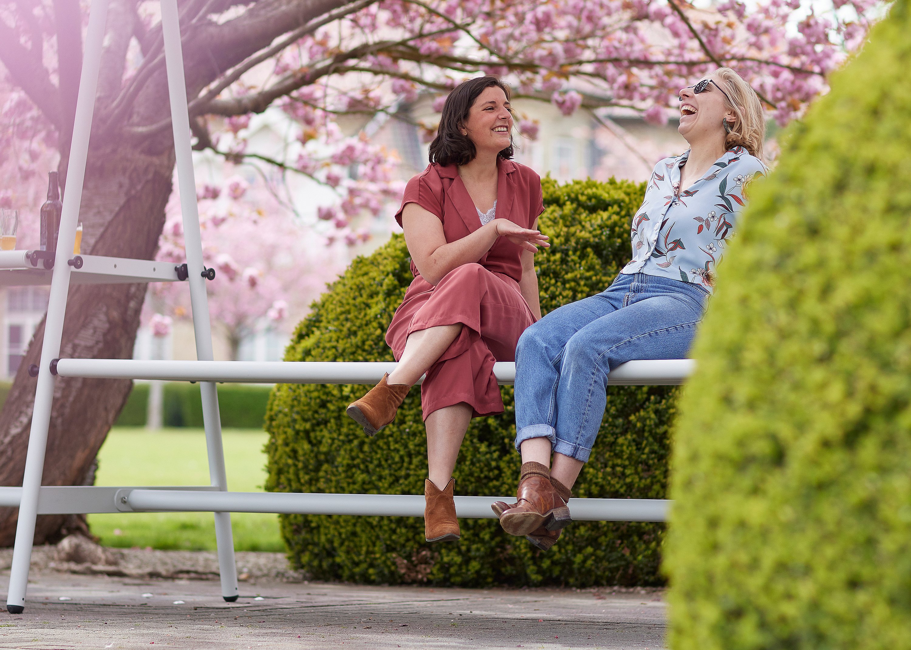 Zwei Frauen sitzen auf einer weißen AMAi-Bank und lachen gemeinsam unter rosa Kirschblüten in einem Garten.