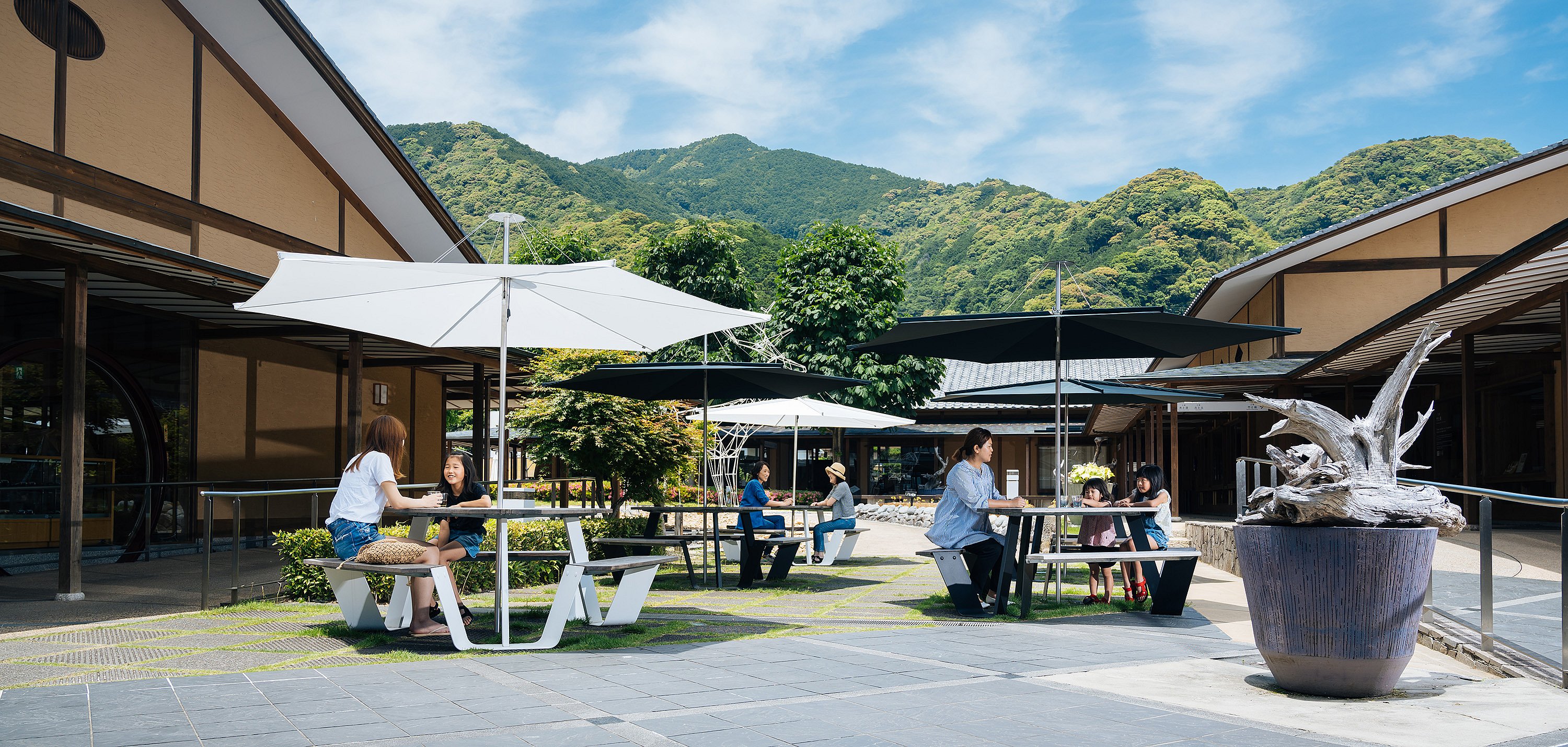 Sitzgelegenheiten im schattigen Garten von Takumishuku mit Gästen, die auf Anker-Sitzmöbeln mit Sonnenschirm sitzen, umrahmt von traditioneller japanischer Architektur und üppigem Grün