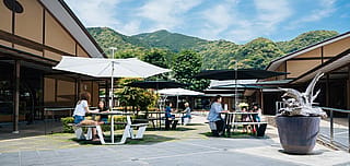 Sitzgelegenheiten im schattigen Garten von Takumishuku mit Gästen, die auf Anker-Sitzmöbeln mit Sonnenschirm sitzen, umrahmt von traditioneller japanischer Architektur und üppigem Grün