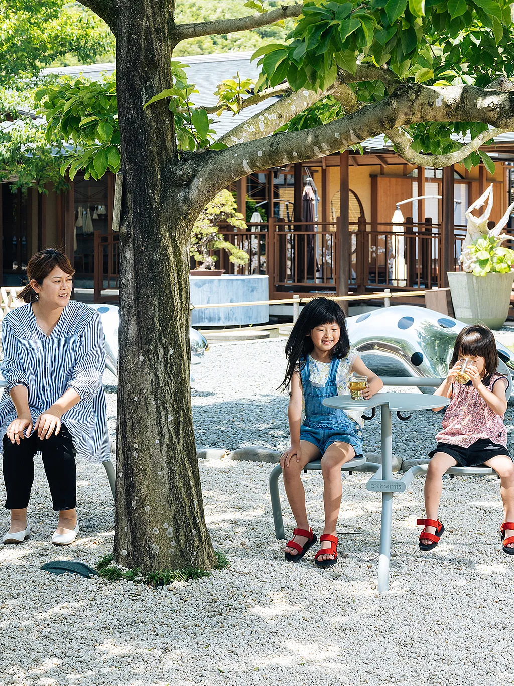 Weitläufiger Außenhof im Takumishuku Arts & Crafts Center in Shizuoka, Japan, mit einem ruhigen Gartenbereich, in dem Bistroo-Duo-Sitzgarnituren auf Steinpflaster stehen, umgeben von grünen Pflanzen und Bäumen