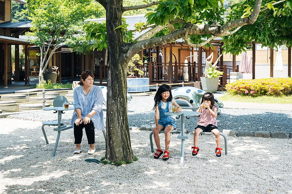 Vue panoramique sur la cour extérieure du centre artistique et artisanal Takumishuku à Shizuoka, au Japon, avec un jardin paisible où sont disposés des ensembles de sièges Bistroo duo sur un dallage en pierre, entourés de plantes vertes et d'arbres.
