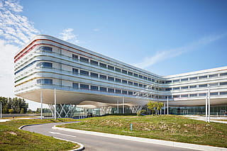 Weitwinkelaufnahme des Klinikgeländes, die das moderne Design des Krankenhauses, die Sitzbereiche im Freien und die landschaftlich gestaltete Umgebung zeigt.