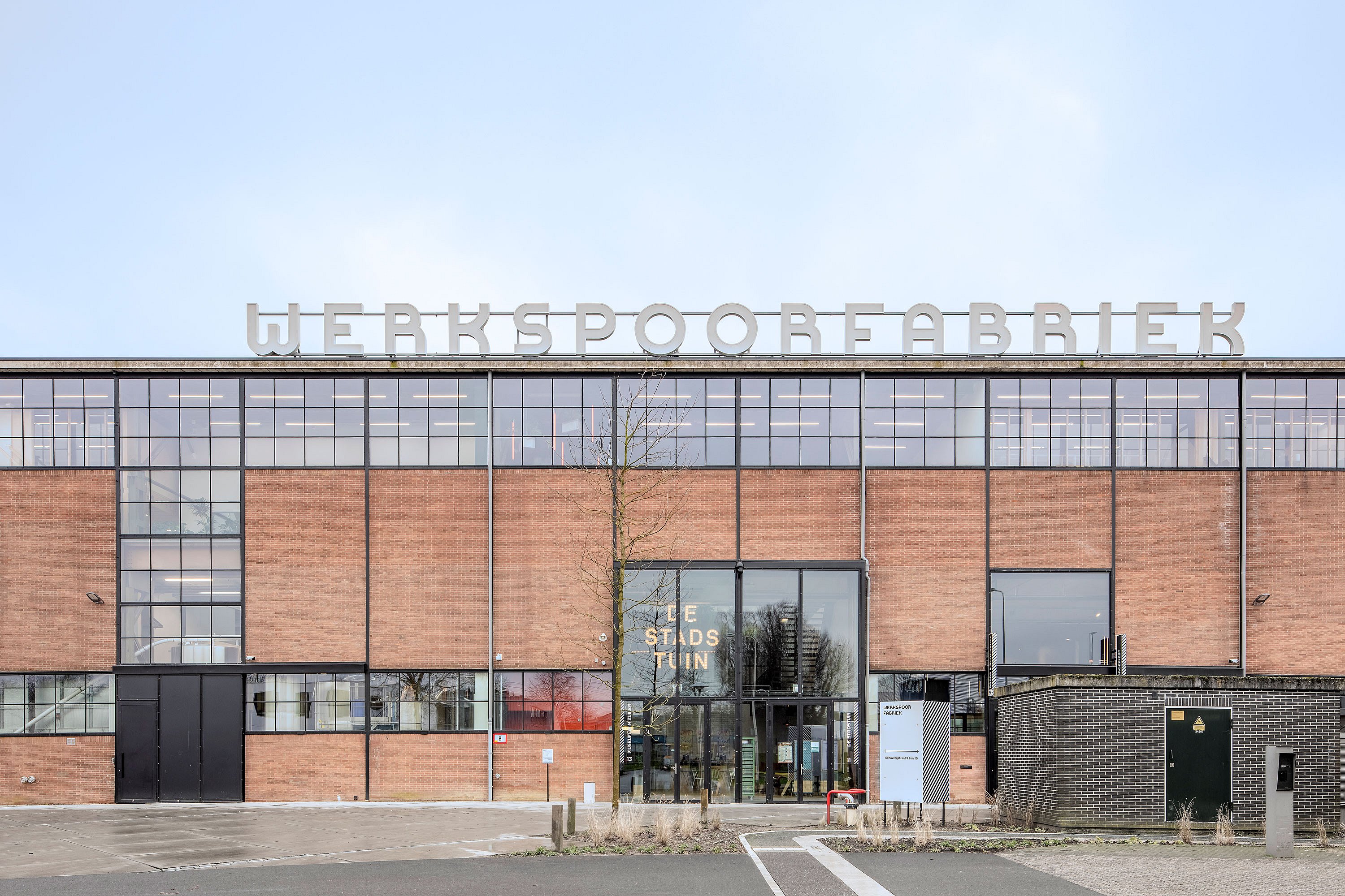   Vue de face du bâtiment en briques de la Werkspoorfabriek, avec ses grandes fenêtres industrielles et son enseigne blanche en gras sur le toit. Cette vue est prise depuis le parvis d'entrée, où l'on aperçoit des portes vitrées et une signalétique au niveau de la rue.