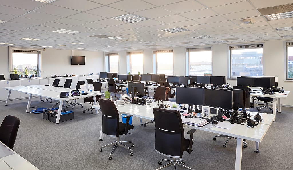 Een open kantoorruimte met rijen houten Marina tafels en banken, waar mensen samen kunnen werken met laptops en documenten.