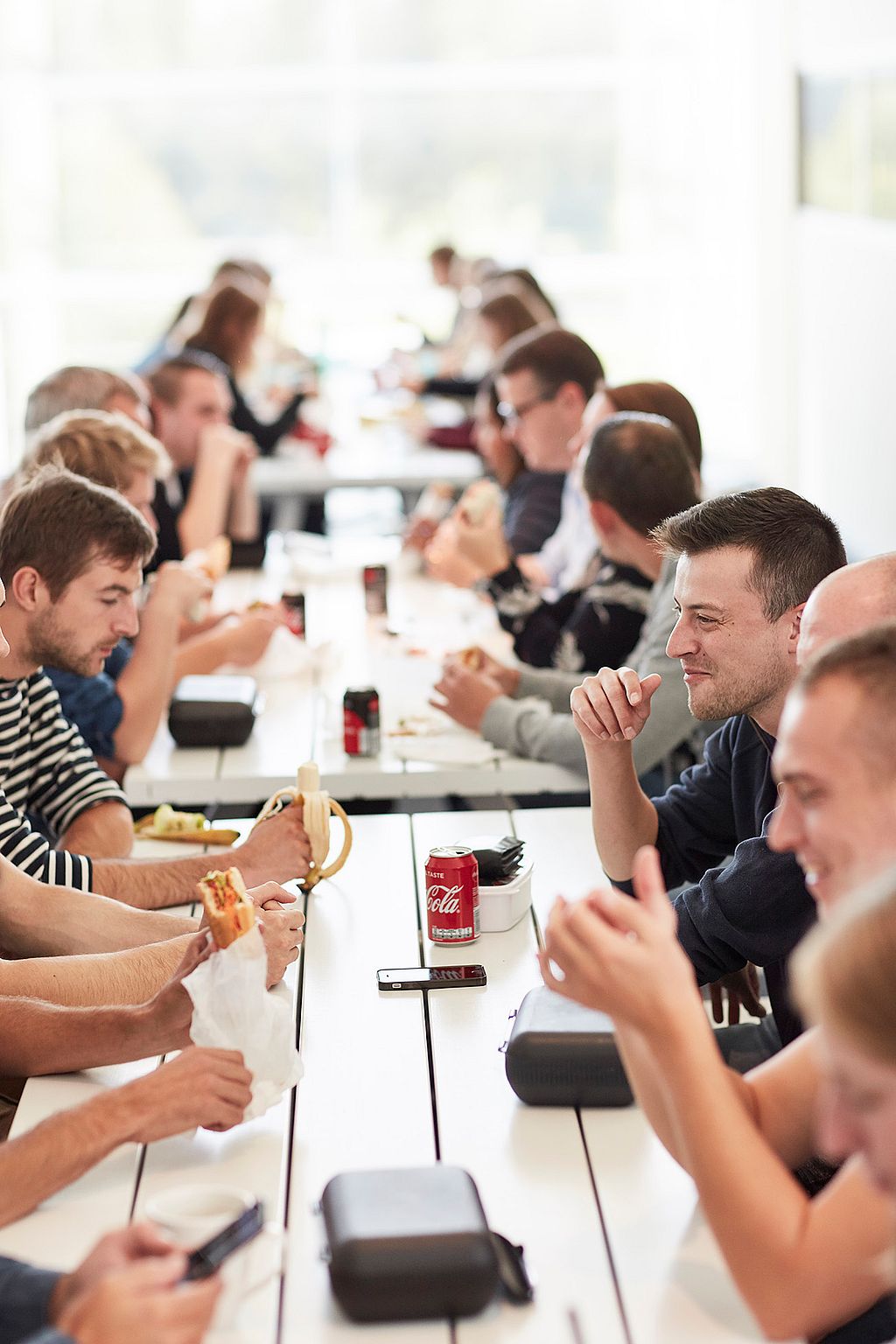 Een groep CCV-collega's zit aan lange witte Marina-tafels in een lichte kantine of kantooromgeving, lunchend en pratend; drankjes, broodjes en persoonlijke spullen staan op de tafels in een levendige, sociale sfeer.