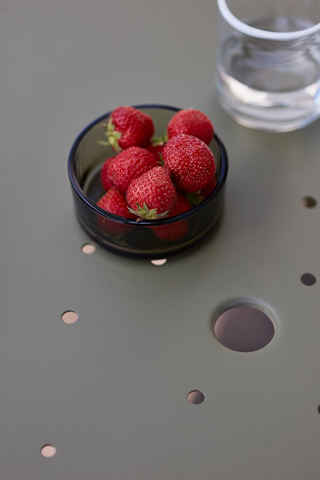 Een schaal met verse aardbeien op een groene Virus-tafel, met een glas water ernaast.