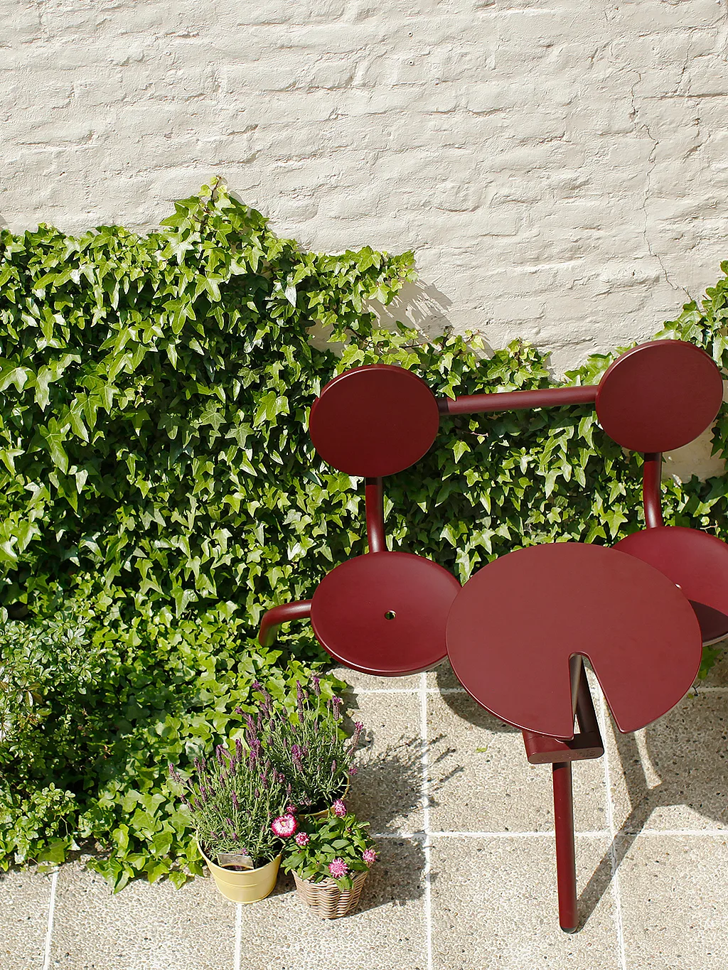 Kleiner urbaner Innenhof mit rotem Bistrotisch und Stühlen an einer mit Kletterpflanzen bewachsenen Mauer – ein gemütlicher, sonniger Essbereich im Freien.