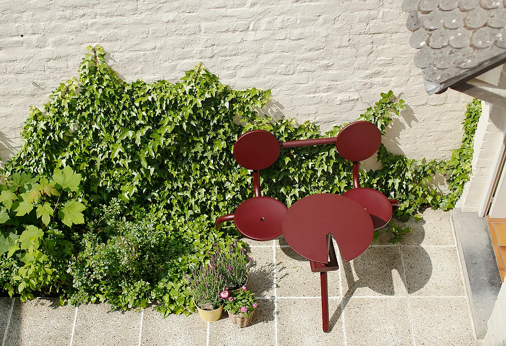 Small urban courtyard terrace with a red Bistroo table and chairs placed against a wall covered in green climbing plants, creating a cozy sunlit outdoor dining spot.