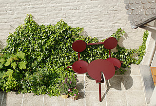 Kleiner urbaner Innenhof mit rotem Bistrotisch und Stühlen an einer mit Kletterpflanzen bewachsenen Mauer – ein gemütlicher, sonniger Essbereich im Freien.