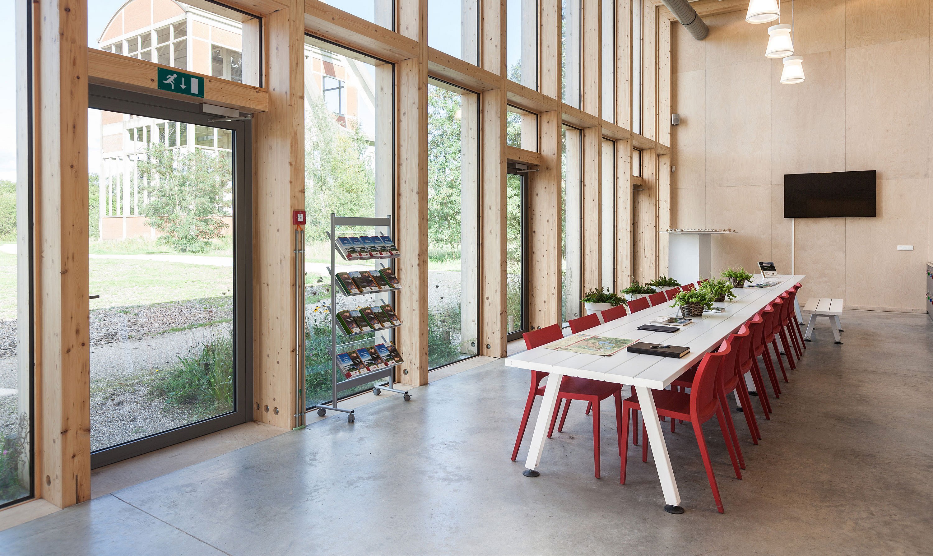   Licht interieur van het bezoekerscentrum met hoge houten ramen die uitkijken op groen, met een lange witte Marina-tafel omringd door rode stoelen, brochures op displaystandaards en potplanten op het tafelblad.