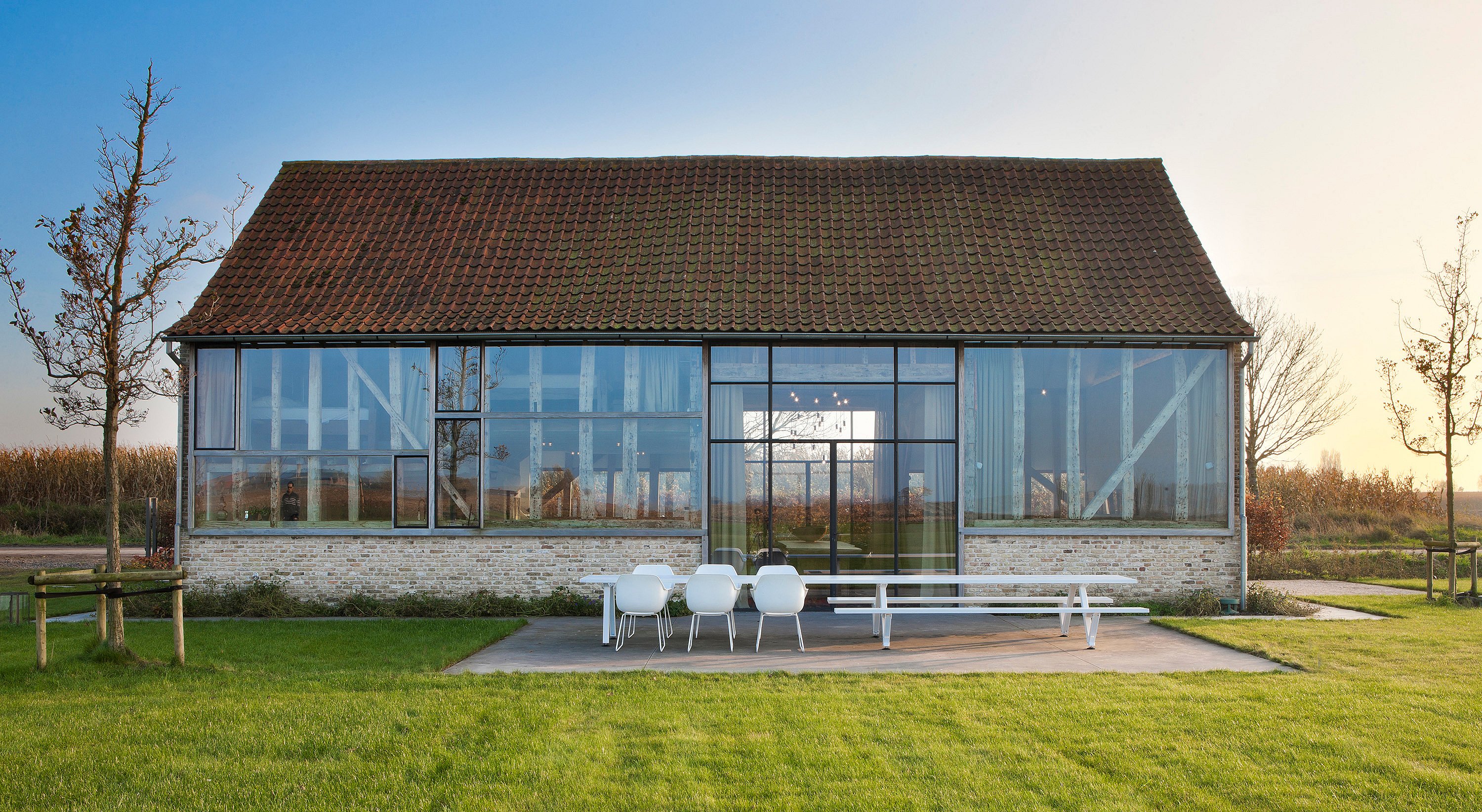 Renoviertes Ferienhaus in einer umgebauten Scheune mit großen Glaswänden, die die originalen Holzbalken freilegen, und einer Terrasse mit Marina-Tisch und Kapitänsstuhl mit Blick auf einen weitläufigen, grünen Garten.