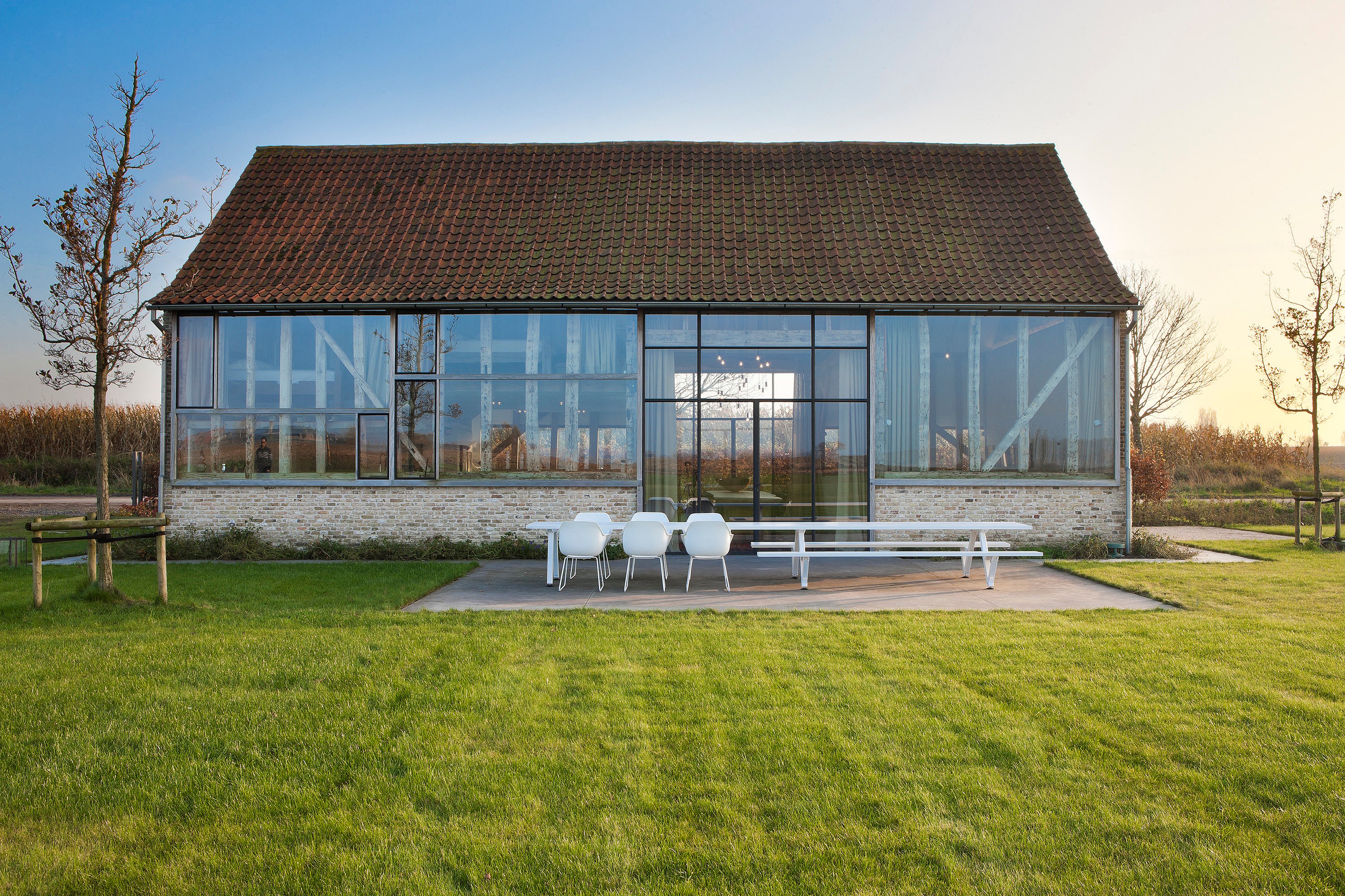 Gerenoveerde vakantiewoning in een schuur met grote glazen wanden die de originele houten balken onthullen en een terras met een Marina-tafel en een Captain's Chair met uitzicht op een ruime groene tuin.