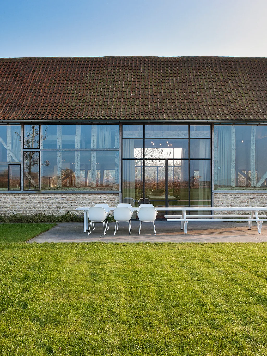 Gerenoveerde vakantiewoning in een schuur met grote glazen wanden die de originele houten balken onthullen en een terras met een Marina-tafel en een Captain's Chair met uitzicht op een ruime groene tuin.