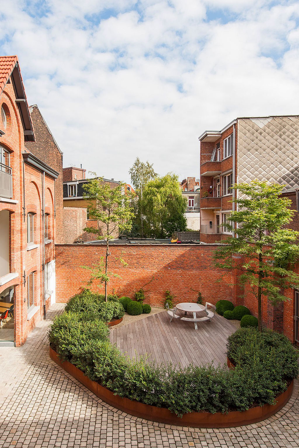 Ein urbaner Innenhofgarten in einem umgebauten Fabrikkomplex mit einem runden Gargantua-Picknicktisch im zentralen, von Grün umgebenen Patio.