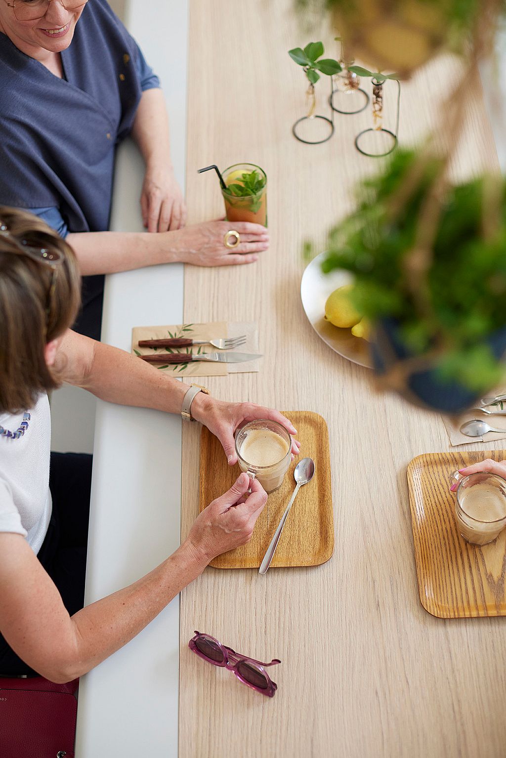 Klanten zitten aan een lange AMAi-tafel met tiki-stoelen in de koffiebar en genieten van een drankje in een ontspannen café-sfeer.