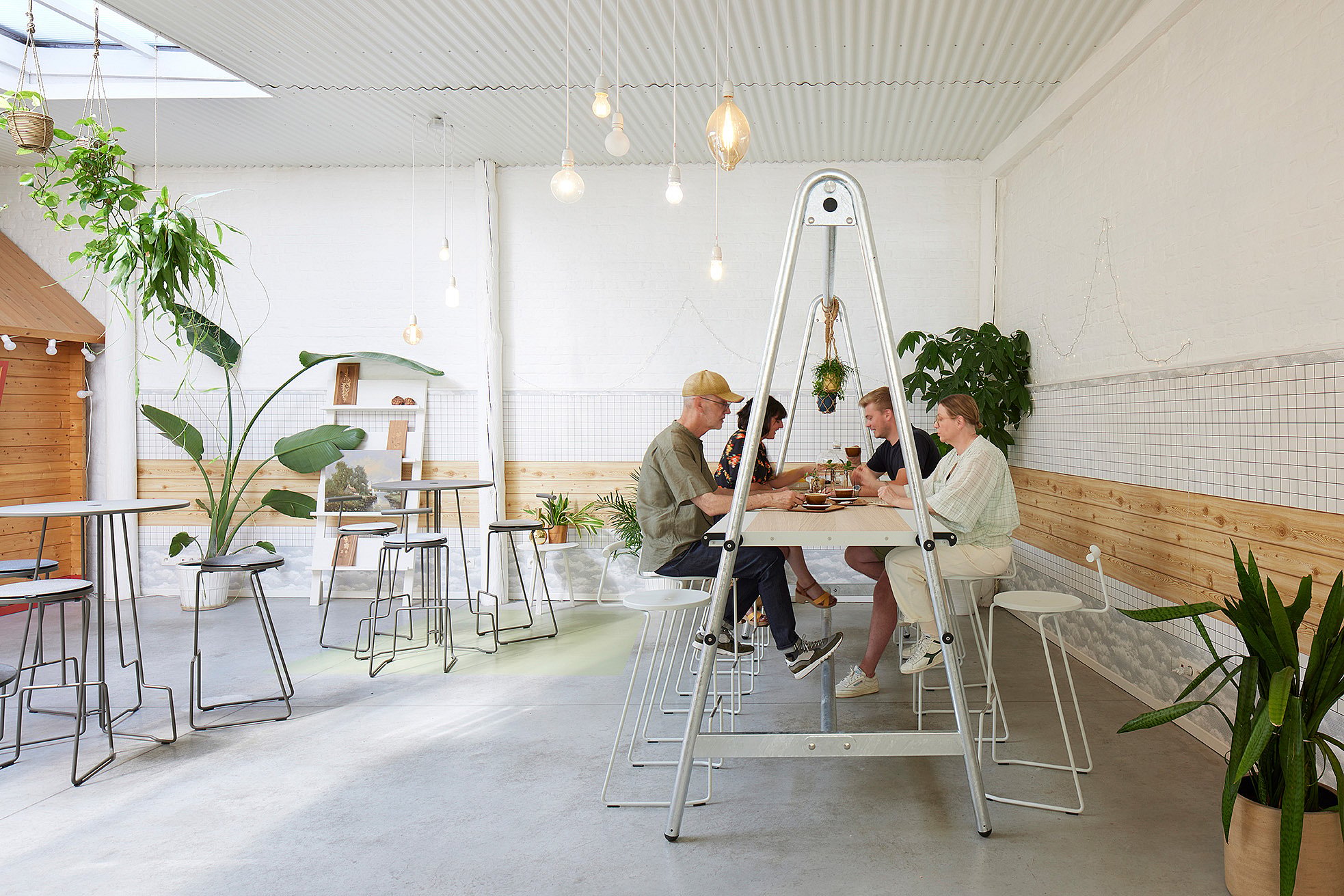 Interieur in industriële stijl van koffiebar Crème de la Crema met een lange AMAi-tafel en tiki-stoelen, omgeven door planten, warme verlichting en klanten die van hun koffie genieten.