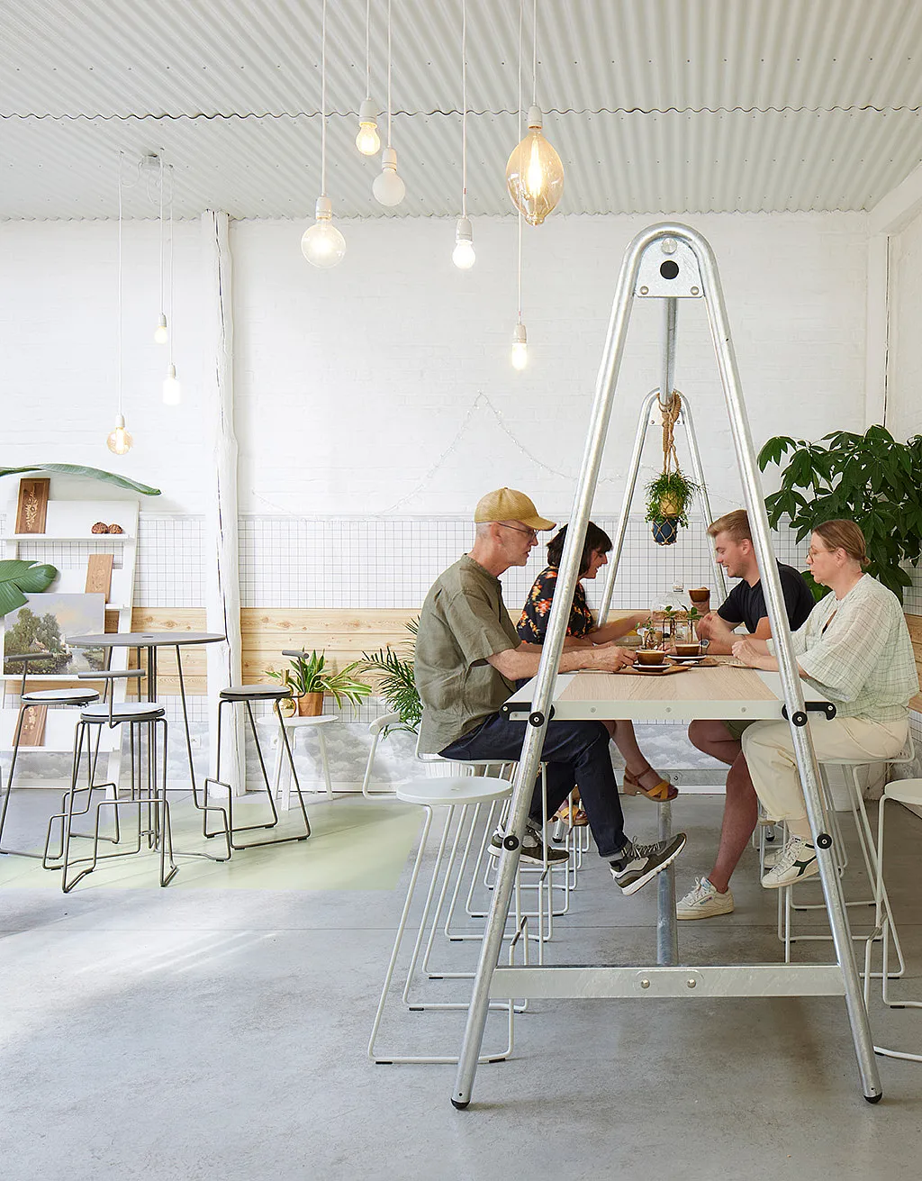 Interieur in industriële stijl van koffiebar Crème de la Crema met een lange AMAi-tafel en tiki-stoelen, omgeven door planten, warme verlichting en klanten die van hun koffie genieten.