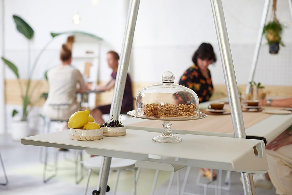 Klanten zitten aan een lange AMAi-tafel met tiki-stoelen in de koffiebar en genieten van een drankje in een ontspannen café-sfeer.