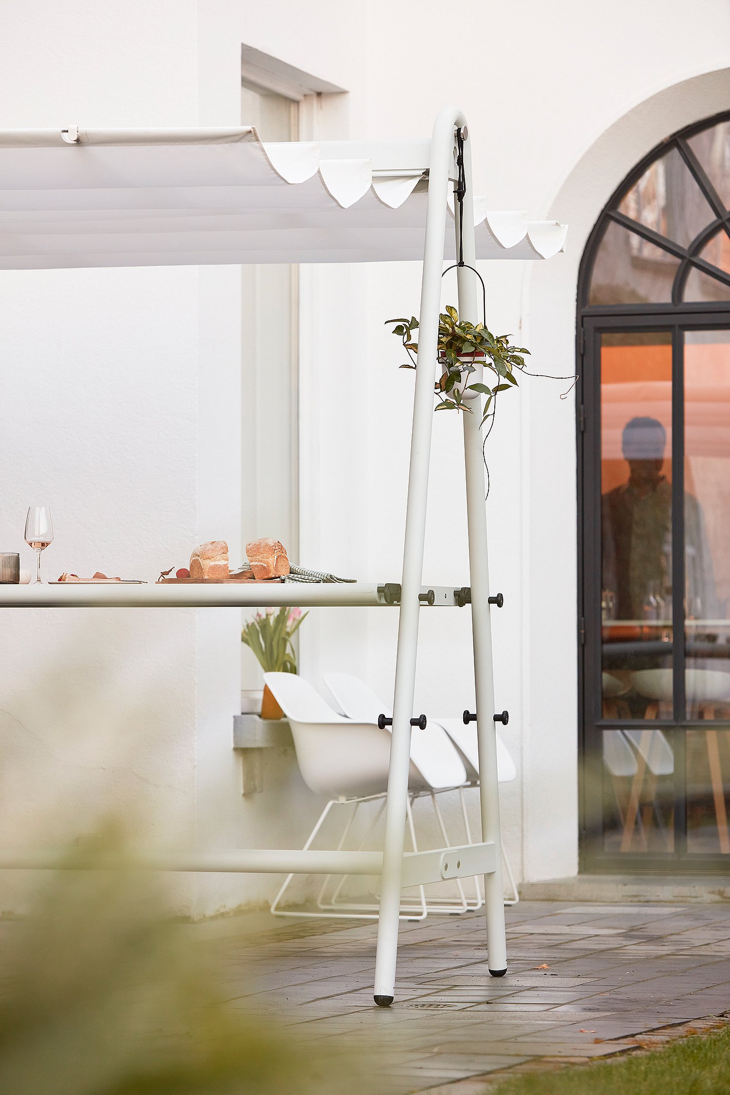 Detail van tafel en stoel: “Close-up van de witte buitentafel met luifel en bijpassende stoel op een terras, met brood en een glas op het tafelblad en de ingang van het huis op de achtergrond. ”