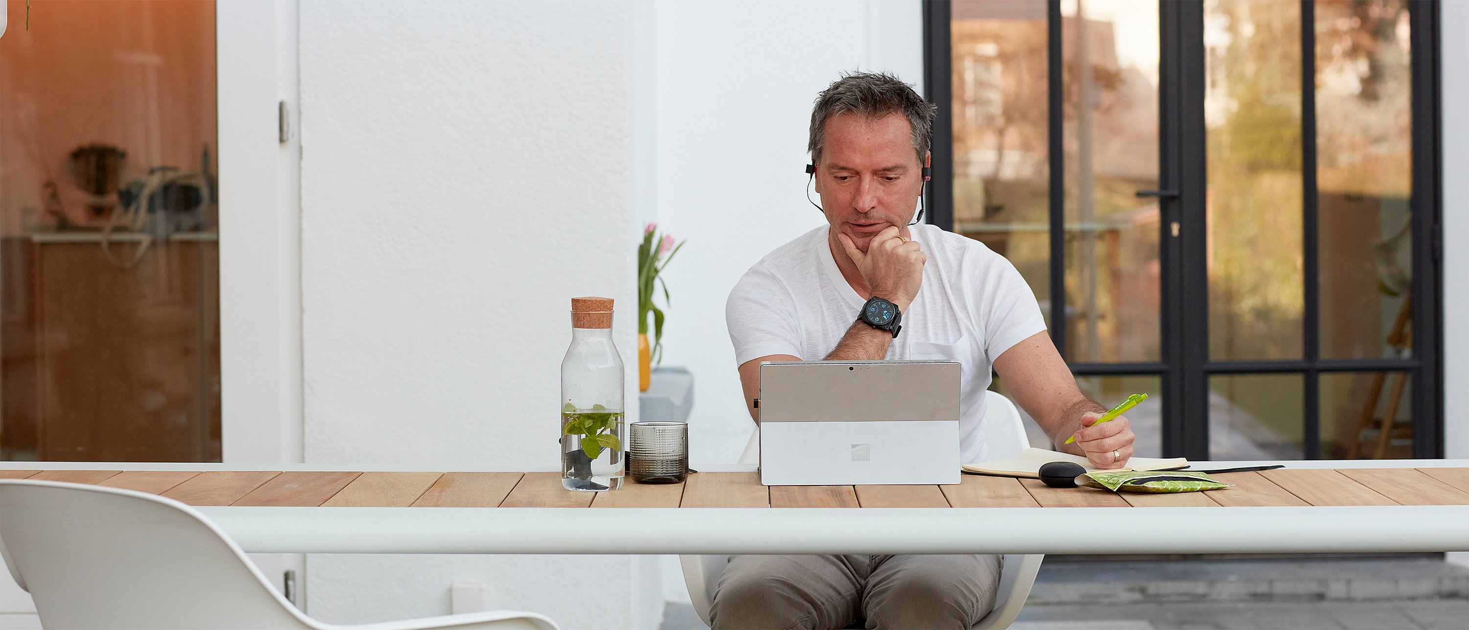Travailler à l’extérieur : " Un homme avec un casque travaille à l’extérieur de la table avec une tablette, un cahier et une boisson, le mobilier servant d’espace de travail en plein air. "