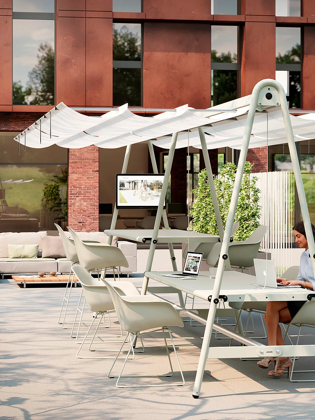 Een buitenkantoor op een terras met een AMAi-tafel en witte Captain's-stoelen onder een geïntegreerd zonnescherm, waar een vrouw op een laptop werkt in een aangelegde binnenplaats naast een modern bakstenen kantoorgebouw.