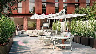 Vue d'ensemble d'une table AMAi blanche avec parasol intégré sur une terrasse arborée devant un bâtiment en briques ; des personnes travaillent et se détendent à proximité.