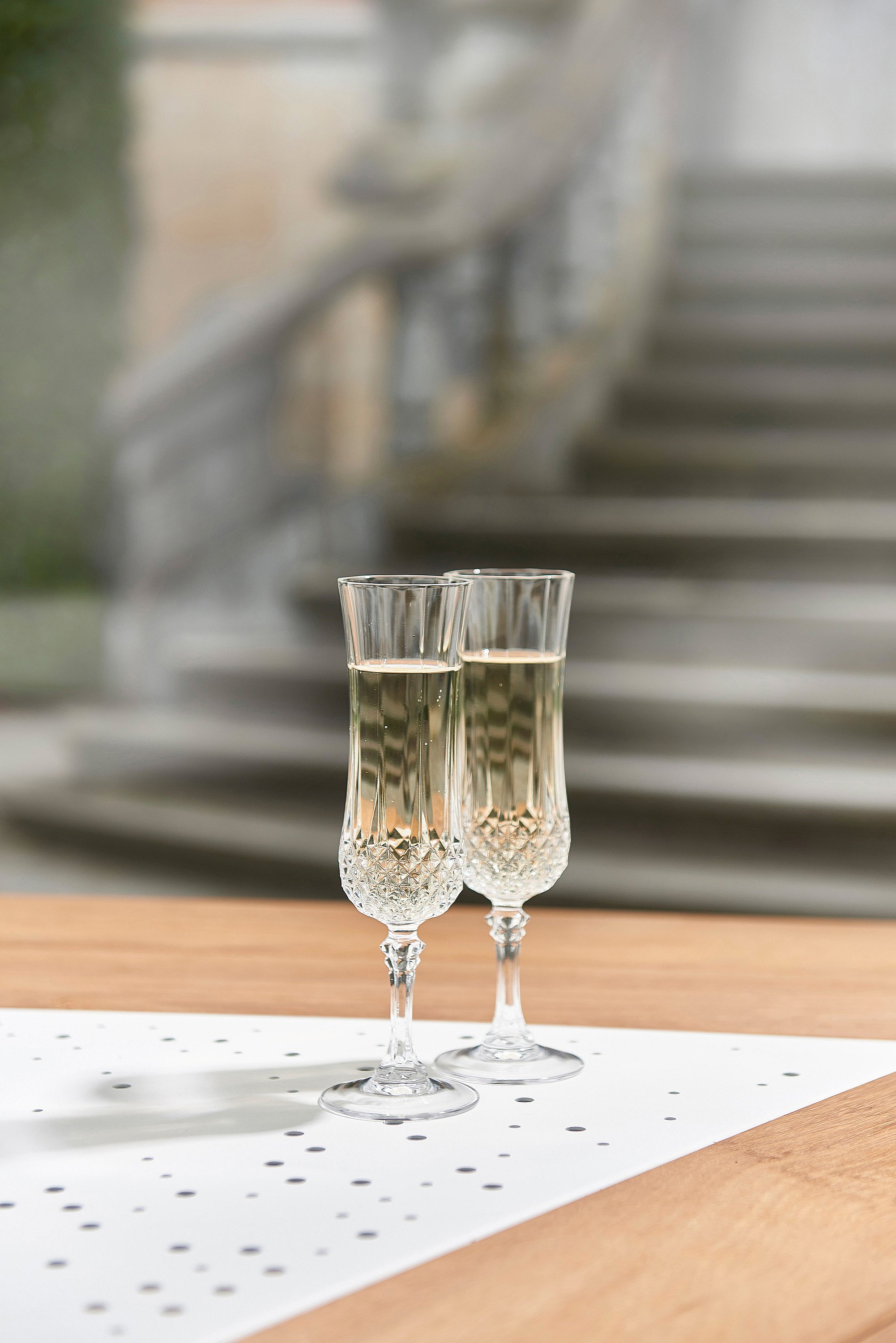   Deux coupes de champagne en cristal remplies de vin pétillant sont posées sur une table de jardin Anker en bois avec incrustations métalliques perforées, avec un escalier en pierre flou et un jardin en arrière-plan.